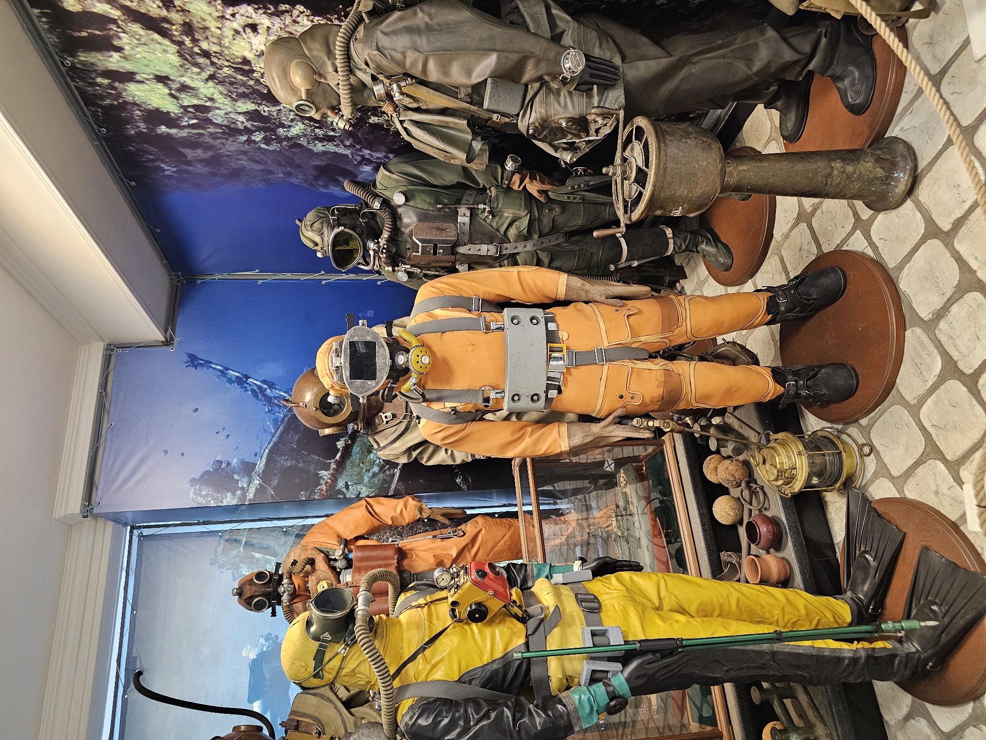 The image shows a display of vintage diving suits arranged vertically. The suits are mounted on circular wooden stands, with a backdrop depicting an underwater scene. From top to bottom, the suits are: one in a dark, possibly leather material; one in olive green; two in orange; and one in bright yellow. All suits have helmets, hoses, and various attachments indicating their function for deep-sea diving. The display appears to be indoors, potentially in a museum or exhibition. The floor is tiled in a diamond pattern. Various diving accessories, such as fins and lamps, are exhibited alongside the suits. It is unknown if the location is within Архангельское, Russia, as there are no visual cues to confirm this.