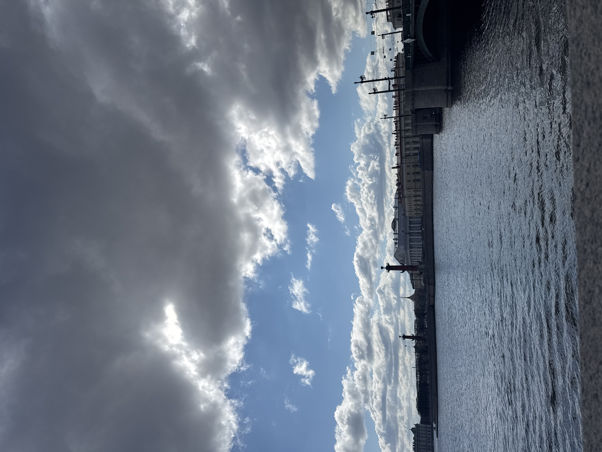 The image shows a section of the Neva River in Saint Petersburg, Russia, under a cloudy sky. The right side of the frame includes a portion of a granite embankment, and the Neva River fills the center. The left side shows the sky filled with varying cloud formations, with patches of blue sky visible in between. Architectural structures line the distant shoreline. The Rostral Columns stand out along the riverbank. A bridge is visible on the upper right.