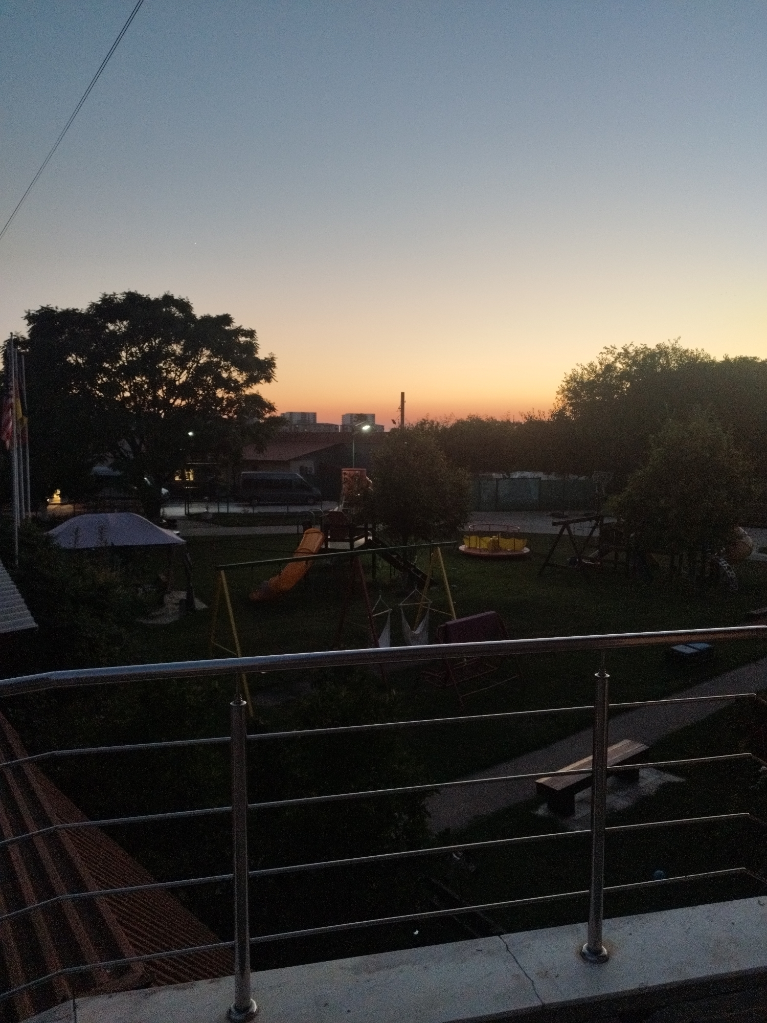 The image shows an outdoor scene at dusk, likely a park or playground area, with a large tree silhouetted against a gradient sky ranging from pale blue at the top to orange and yellow near the horizon. Several playground structures are visible, including a slide, swings, and a merry-go-round. A few buildings and what appears to be a van or bus are in the background. In the foreground, a metal railing overlooks the scene, suggesting the vantage point is from a balcony or elevated walkway. There are no people visible in the frame. The overall impression is one of a quiet evening in a residential or recreational area.