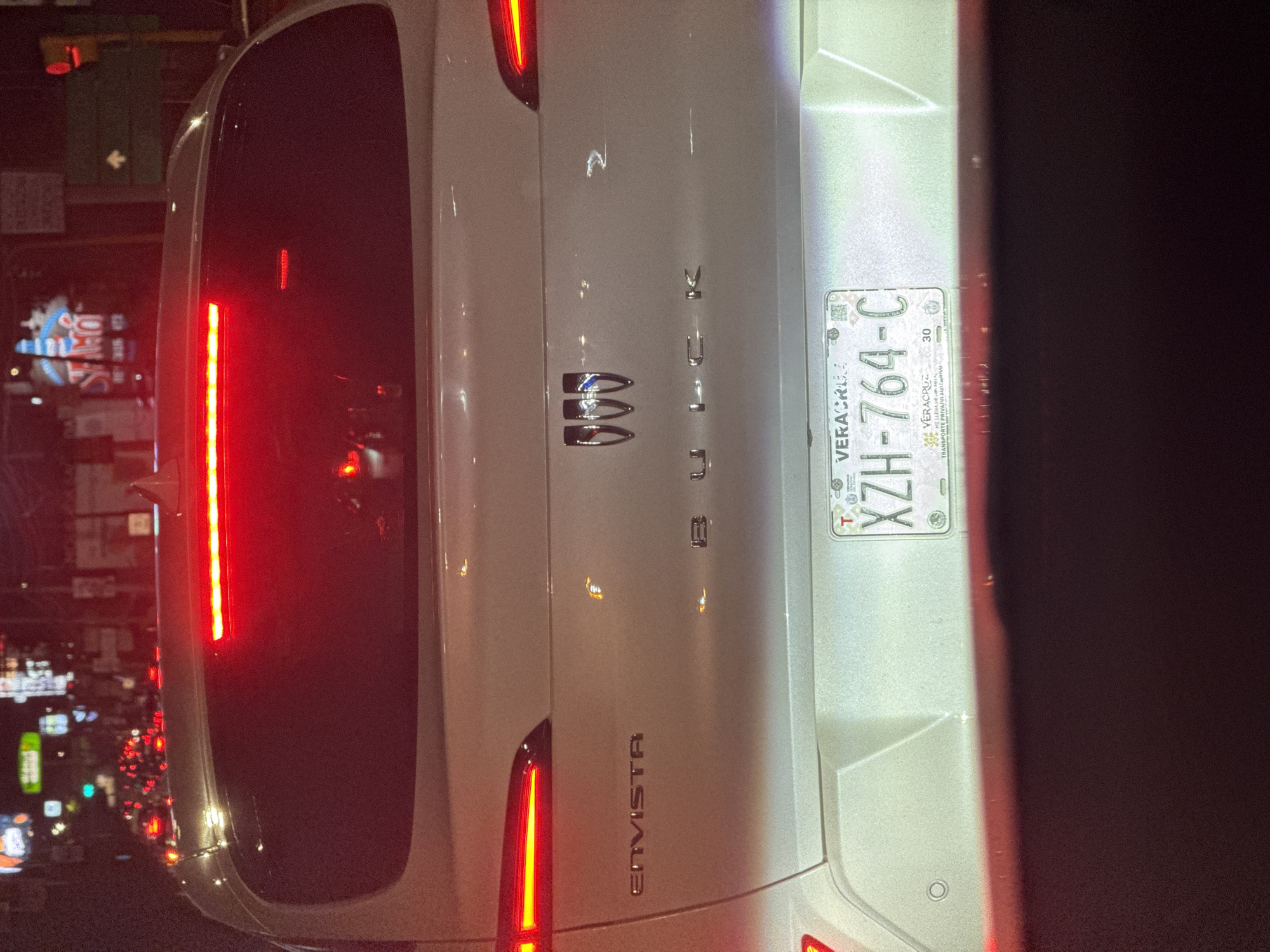 The image captures the rear of a white Buick Envista SUV, illuminated at night. The car is positioned vertically in the frame, with its red taillights prominently lit. The "ENVISTA" model name is visible on the lower left of the trunk, while the "BUICK" brand name and its three-shield emblem are centered.
A Mexican license plate is affixed to the right side of the trunk. The plate clearly displays "VERACRUZ" at the top, followed by the alphanumeric sequence "XZH-764-C". Below the main number, smaller text indicates "TRANSPORTE PRIVADO MERCANTIL" and the number "30".
The background is dark and blurry, typical of a nighttime urban environment. Numerous out-of-focus lights, likely from other vehicles' taillights, streetlights, and distant illuminated signs, suggest the car is on a city street, possibly in traffic. The scene conveys a sense of a busy night in an urban setting. While the car's license plate is from Veracruz, the given context specifies the location as Puebla, Mexico, suggesting the image was captured in a Mexican city at night.