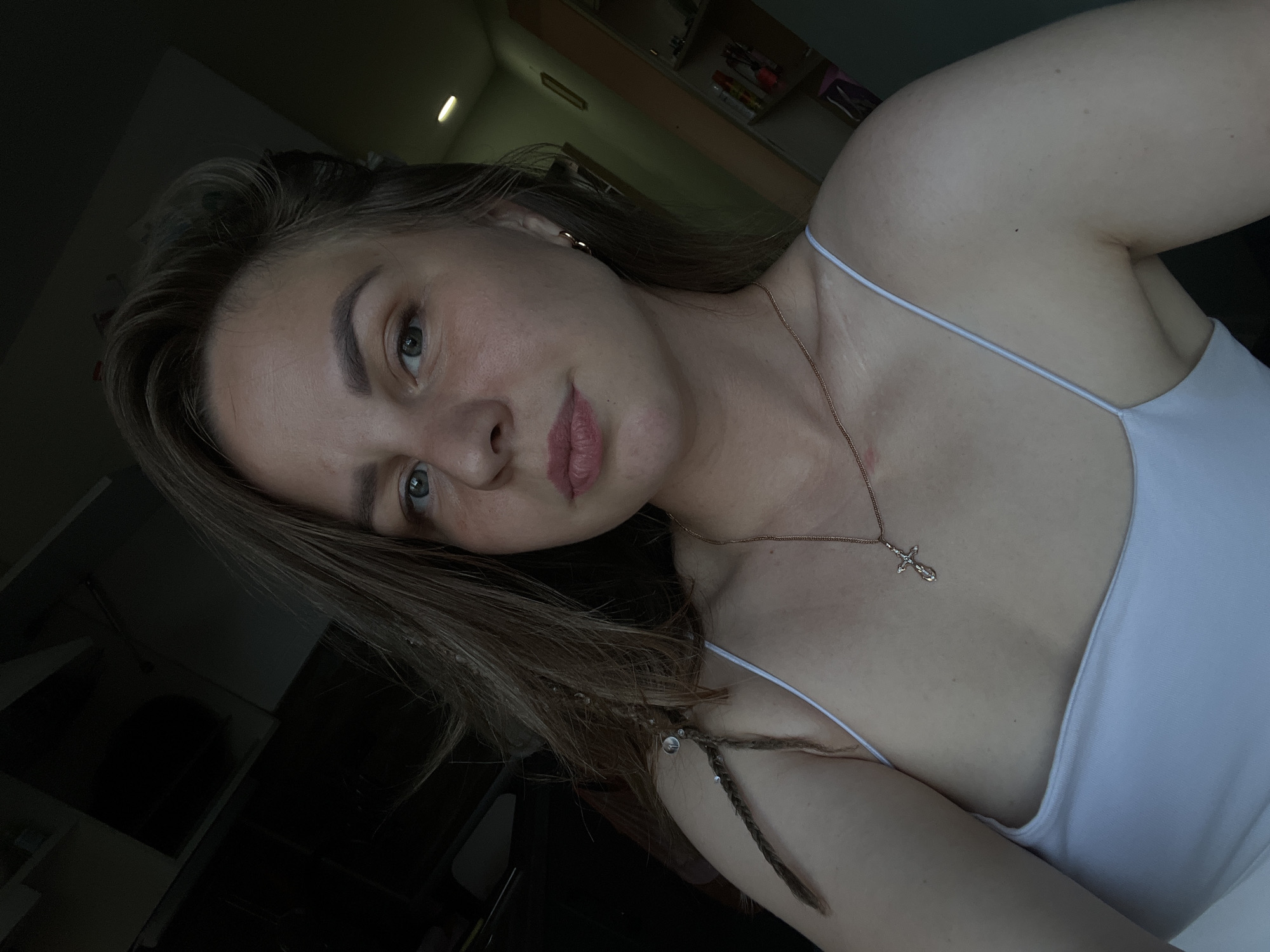 A young woman is featured in a close-up selfie, captured indoors in what appears to be a room in Izhevsk, Russia. She has light skin, brown hair, and green or hazel eyes, looking directly into the camera with a neutral to slightly smiling expression. Her lips are adorned with pink lipstick.
She is wearing a white spaghetti strap top or camisole, revealing her bare shoulders and upper chest. Around her neck, she wears a thin gold chain with a small, ornate gold cross pendant. Her brown hair is styled with several small braids, some of which are decorated with tiny silver-colored beads or rings near her shoulder. A small, heart-shaped gold earring is visible on her left ear.
The image is taken at a tilted angle, with natural light illuminating her face and upper body from the left. The background is mostly dark and out of focus, though hints of shelves with objects and a light fixture on a wall or ceiling can be discerned, suggesting a domestic setting. No specific weather conditions are visible. There is no discernible text in the image.