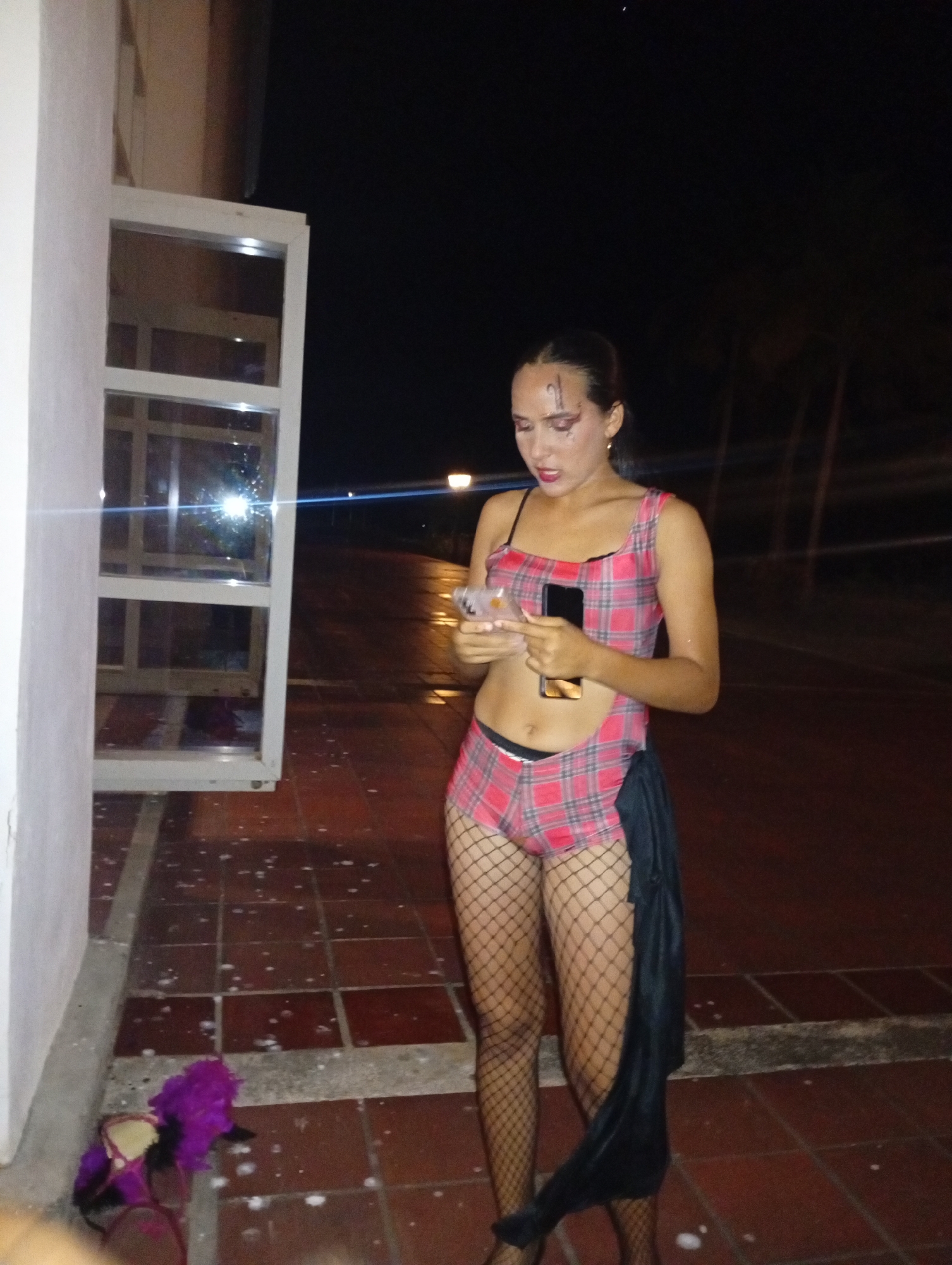 A female stands outside near a white building with a window. She is wearing a red plaid outfit with black fishnet stockings and holding a phone and a clear plastic cup. On the ground near her is a purple and black object. The setting is dark, possibly nighttime, with a lit street visible in the background, indicating a location in Pedro González, Venezuela. The ground is tiled.