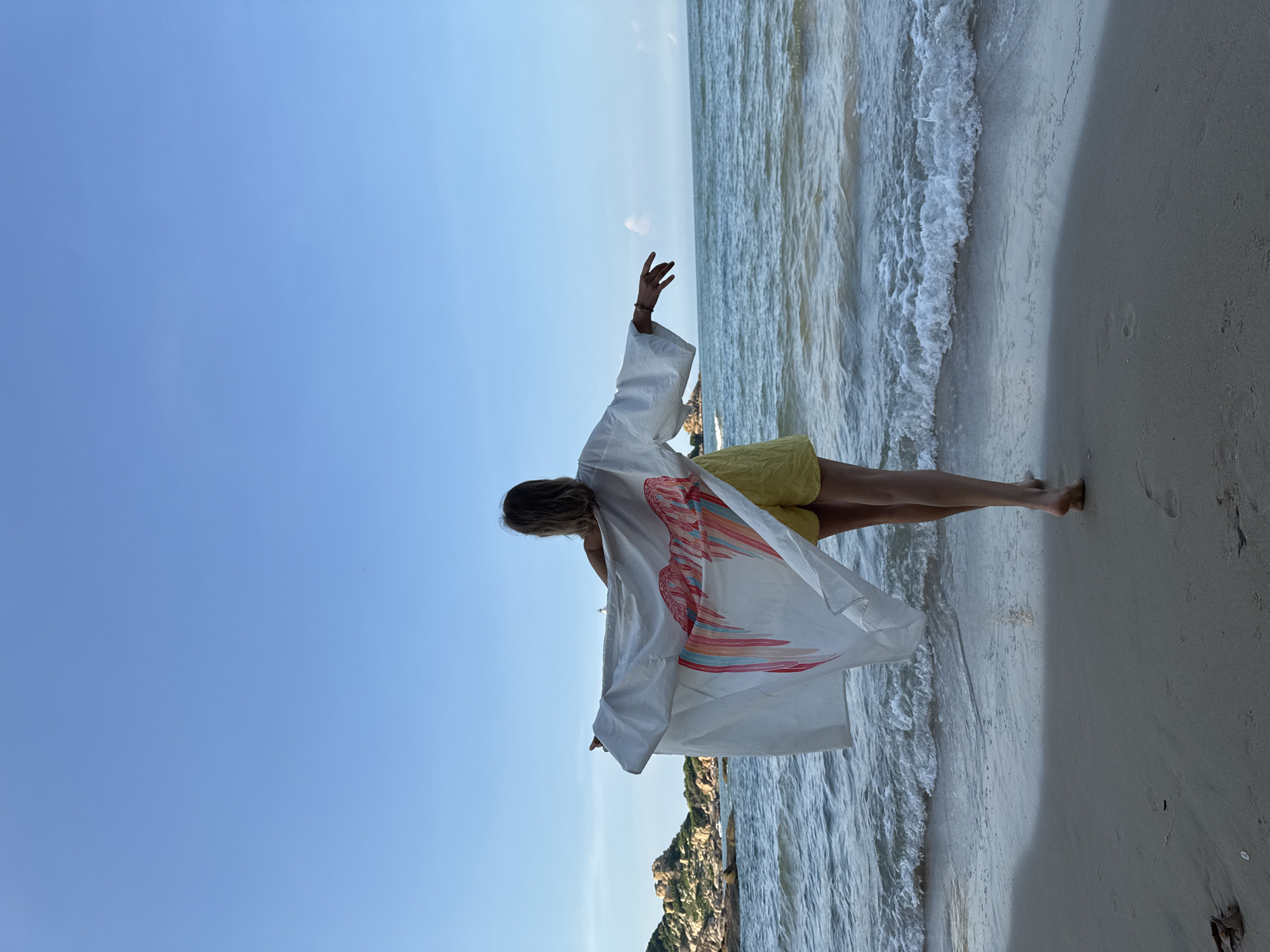In the city of Thuận Thành, Vietnam, a lone woman stands on a sandy beach, facing away from the camera. She wears a white open-fronted garment with a colorful wing-like design on the back and yellow shorts. Her arms are outstretched, holding the garment out to her sides as if to embrace the scene. Gentle waves lap at the shore in front of her. In the distance, a rocky outcrop rises from the sea under a clear blue sky. The scene appears to be captured during the late afternoon, with the sun casting a warm glow on the water and sand.
