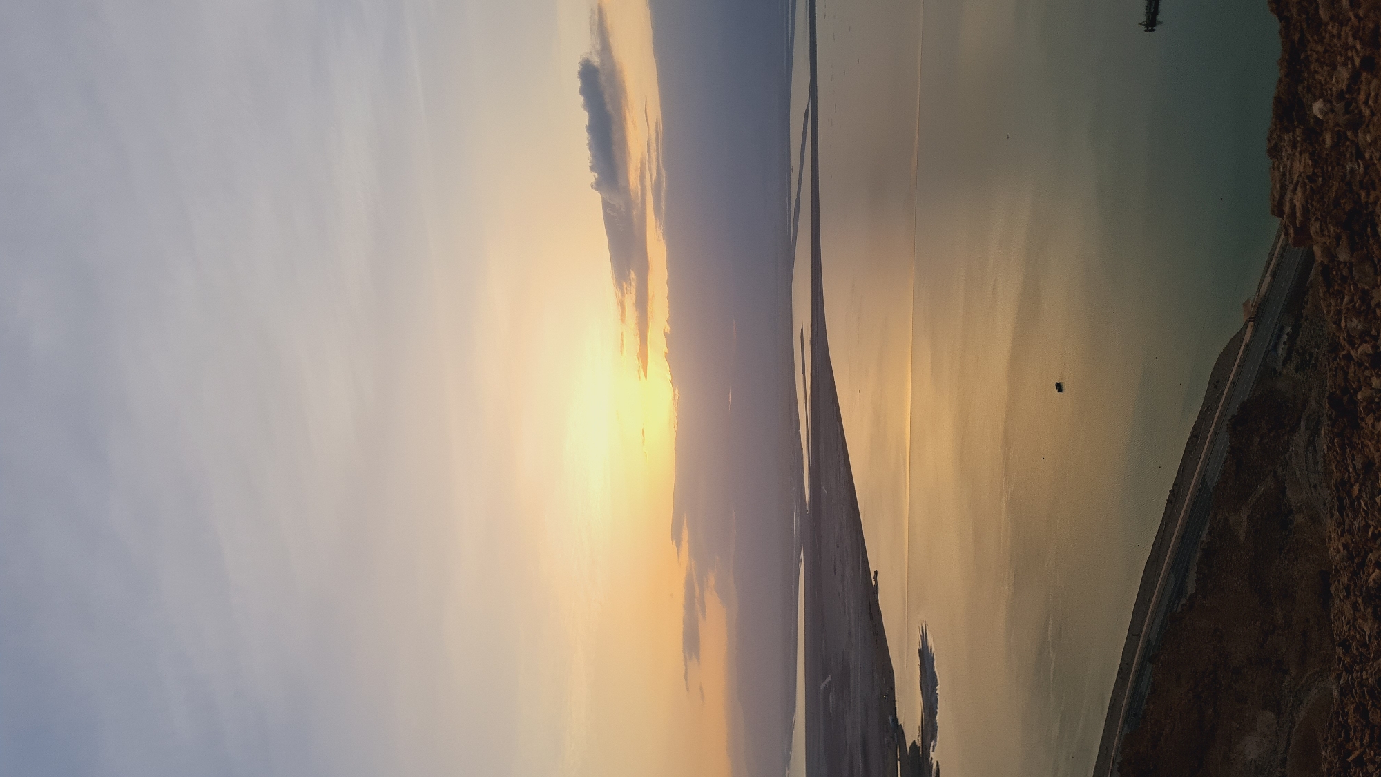 The image depicts a sunset over the Dead Sea, located in the Tamar Regional Council, Israel. The sky shows shades of blue and orange with cloud formations near the horizon. The sun's light reflects off the water's surface, creating a golden hue. A strip of land divides the water body into two sections. The right side of the image features rocky terrain alongside a road. In the water, there is a small, dark square, which is most likely a distant object.