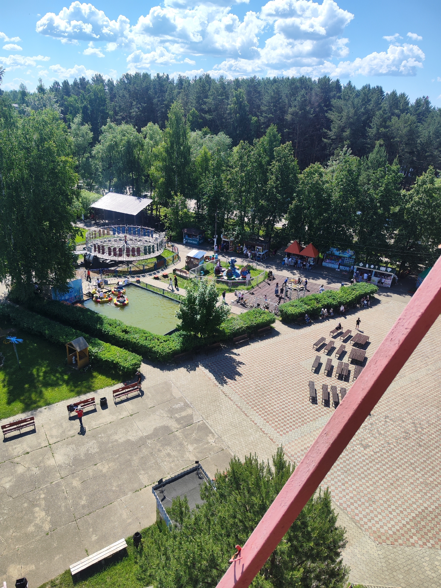 The image provides an aerial view of an amusement park on a sunny day with a partly cloudy sky. In the foreground, a rusty red railing runs diagonally across the bottom right of the frame. The middle ground of the park features a small pond with several yellow bumper boats carrying occupants, surrounded by green hedges. Adjacent to the pond is a spinning amusement ride with a circular platform and gondolas. Various food stalls and smaller attractions are visible further into the park, with people milling about. The background is dominated by a dense forest of tall green trees. In the lower left section, a paved area has several empty park benches and trash cans, with some patches of grass. A section of patterned brick paving is visible in the lower right quadrant, also with benches.