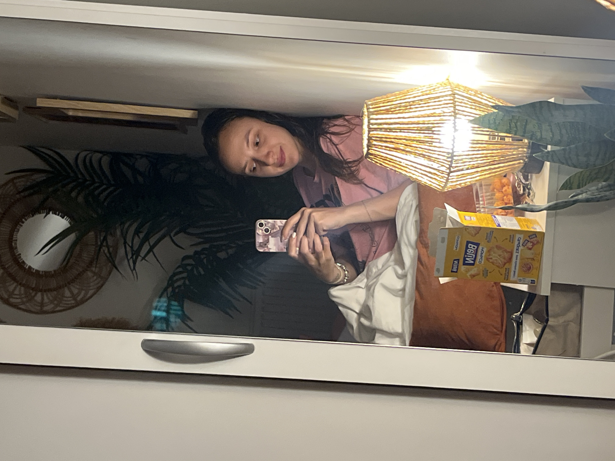 The image is a mirror selfie capturing a young woman in a room, holding her smartphone. She has long, dark hair and is wearing a pink top, with a bracelet visible on her wrist. Her expression is a soft, relaxed smile as she looks towards the phone, which has a light-colored, patterned case.
The reflection reveals a cozy interior. On the left, a large, dark green, palm-like plant stands prominently, partially obscuring a circular decorative mirror with a woven frame. Above the plant, wooden shelves are visible. To the right, a square-shaped lamp with a woven, straw-like shade emits a bright, warm light. Next to the lamp, an open yellow box of snacks, identified as "КУКУРУЗНЫЕ ЧИПСЫ" (Corn Chips) from the visible text, and a clear container with orange, round snack items are placed on a surface. Another green plant, possibly a snake plant, is partially seen near the snacks.
Elements of a bed or couch, including a rust-colored pillow and a white blanket, are visible. The bottom edge of the mirror appears to be part of a white piece of furniture with a silver handle. The warm lighting from the lamp suggests it might be evening or indoors. The scene is located in Vitry-sur-Seine, France.