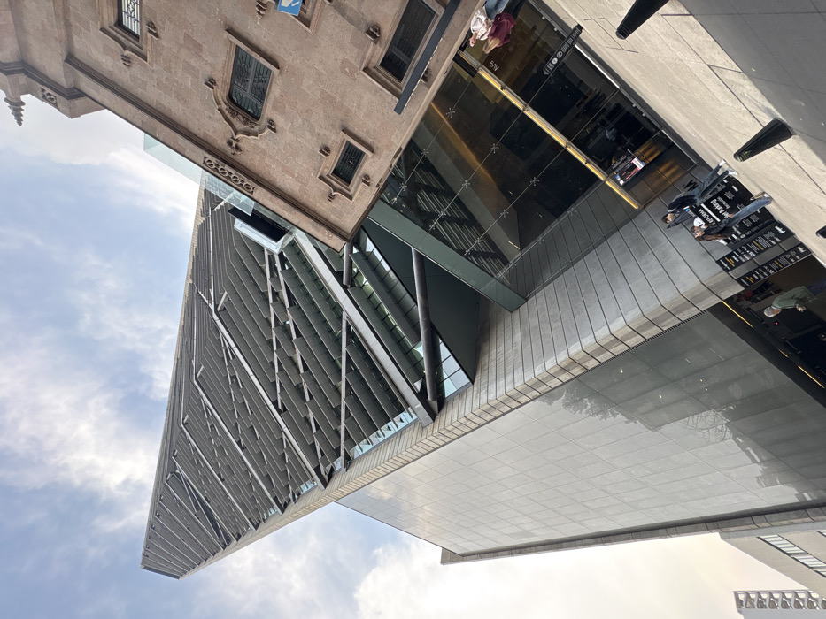 The image presents a low-angle, tilted view of the Torre Reforma skyscraper and an adjacent historic building on Paseo de la Reforma in Mexico City, Mexico. The skyscraper, a tall, slender structure, occupies the left and central portion of the frame, characterized by its modern design featuring an external facade composed of a prominent diagonal grid of grey louvers or fins, with dark glass panels below. Its lower sections include large reflective glass surfaces and a lighter, textured stone or concrete base. To the right, an older, multi-story building made of reddish-brown stone exhibits traditional architectural elements, including arched and rectangular windows, decorative stone carvings, and a pointed spire-like finial at its uppermost visible section. A modern glass and steel canopy extends from the skyscraper's base towards the historic structure, physically connecting them. The sky overhead is partly cloudy. At street level, three pedestrians are visible: one male in a green jacket near the bottom left, and a male and female standing near a black informational sign displaying text that includes "Reforma" and "Parking." Several dark, conical bollards are positioned along the sidewalk.