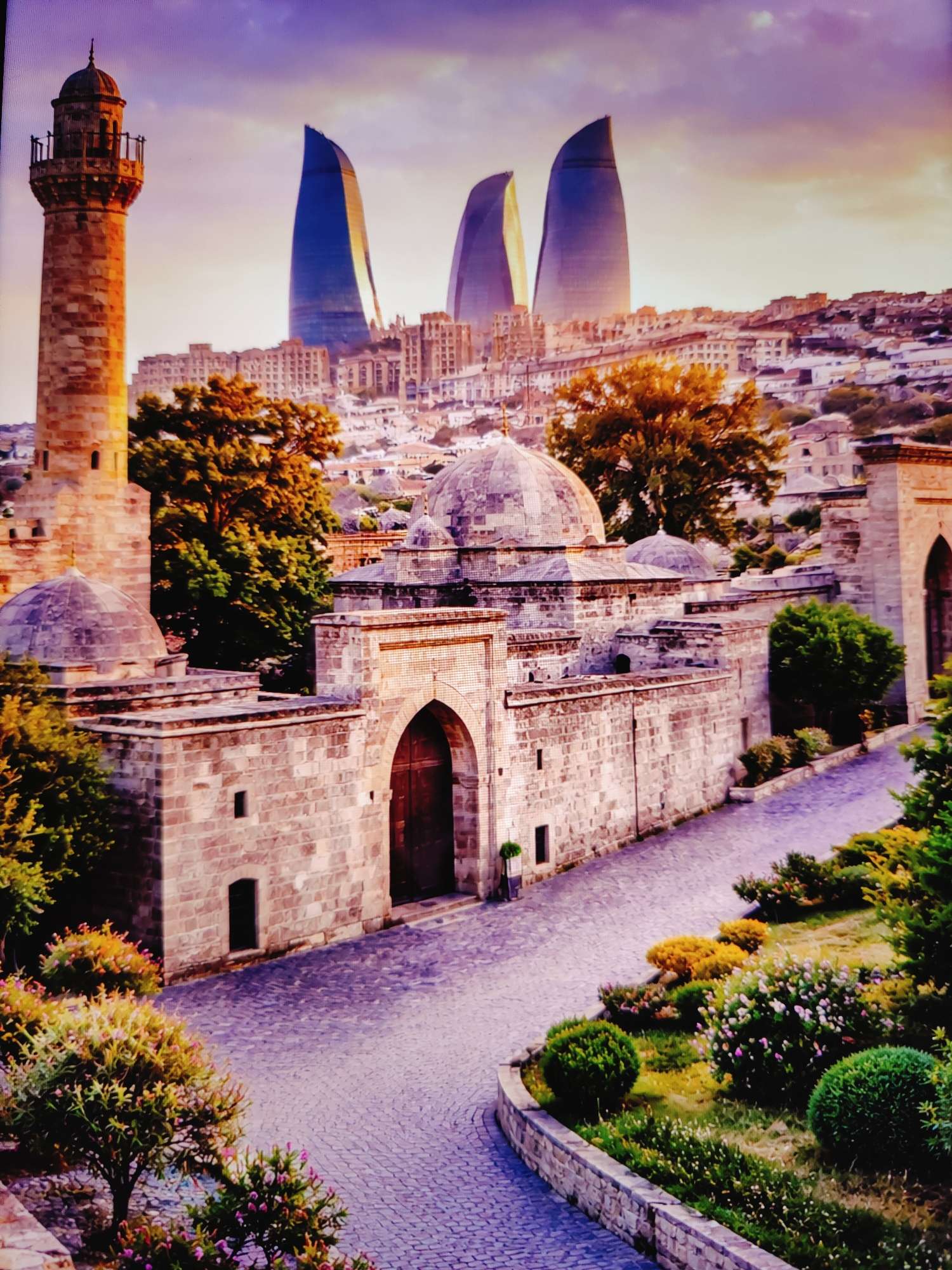 The image depicts a picturesque view of the Old City (Icherisheher) in Baku, Azerbaijan, at dusk. In the foreground, a stone minaret, part of an ancient mosque or religious complex, stands tall, its warm, sun-kissed stone contrasting with the dusky sky. Adjacent to the minaret are stone walls and domes of historical buildings, characterized by their weathered stone construction and intricate architectural details. A paved pathway, made of cobblestones, curves through the scene, lined with lush greenery and flowering bushes.
In the background, the modern Flame Towers, iconic skyscrapers of Baku, rise majestically against the twilight sky, their flame-like silhouettes illuminated and reflecting the colors of the setting sun and the approaching night. The juxtaposition of ancient architecture in the foreground and contemporary skyscrapers in the background highlights the unique blend of history and modernity that defines Baku. The sky is painted with hues of purple, orange, and yellow, suggesting a dramatic sunset. There are no people visible in the scene. The overall atmosphere is serene and awe-inspiring, capturing the beauty of Baku's historic heart with its modern marvels in the distance.