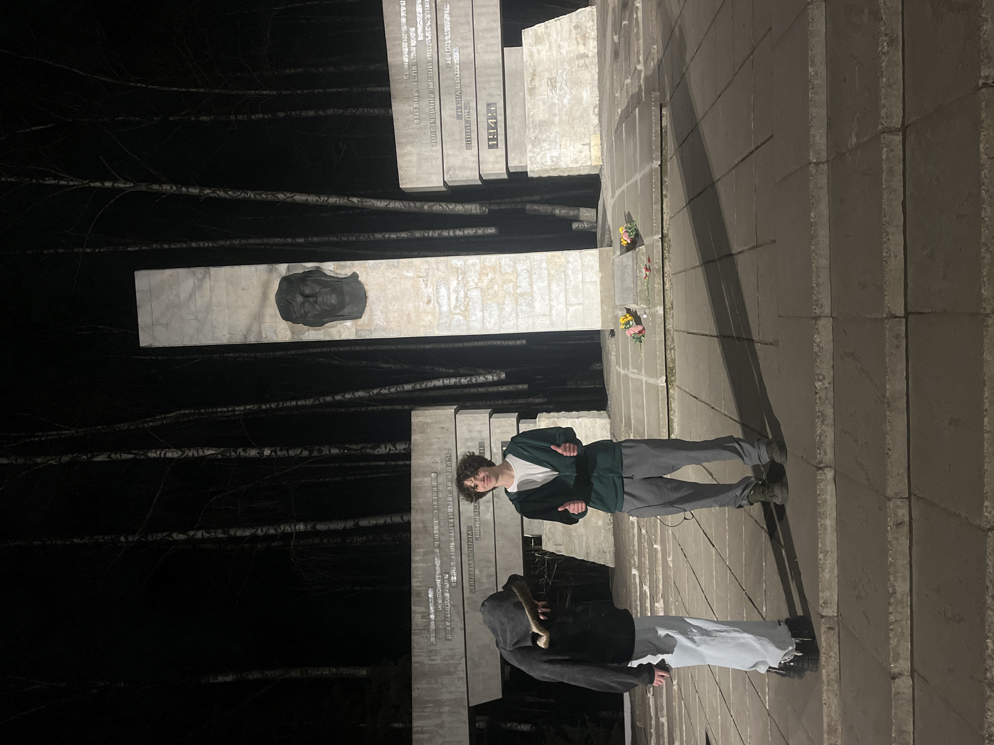 Two individuals stand at a World War II memorial located in Saint Petersburg, Russia. The memorial consists of several stone pillars inscribed with text and a sculpture of a person's face. Flowers are placed at the base of the memorial. The surrounding area includes trees and a paved walkway. One person is wearing a green jacket and grey pants, while the other is wearing a dark hoodie and light-colored pants.