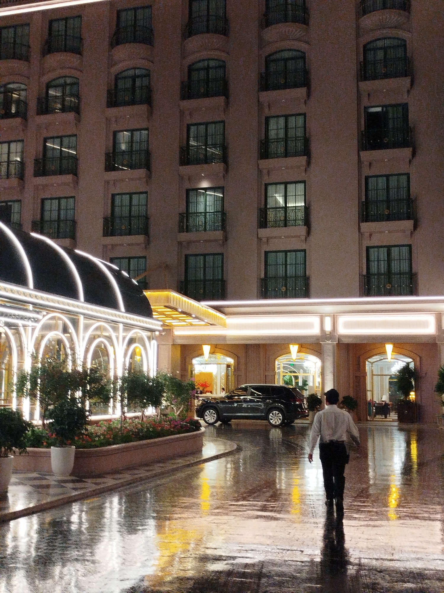 An exterior shot shows a multi-story building at night in Lâm Viên - Đà Lạt, Vietnam. The building is made of a light-colored material and has multiple rows of windows with small balconies. An ornate, brightly lit canopy leads into the building's entrance. A row of plants and flowers separates the canopy from the foreground. A man walks away from the camera on a wet, reflective surface. Two dark-colored SUVs are parked under the canopy.