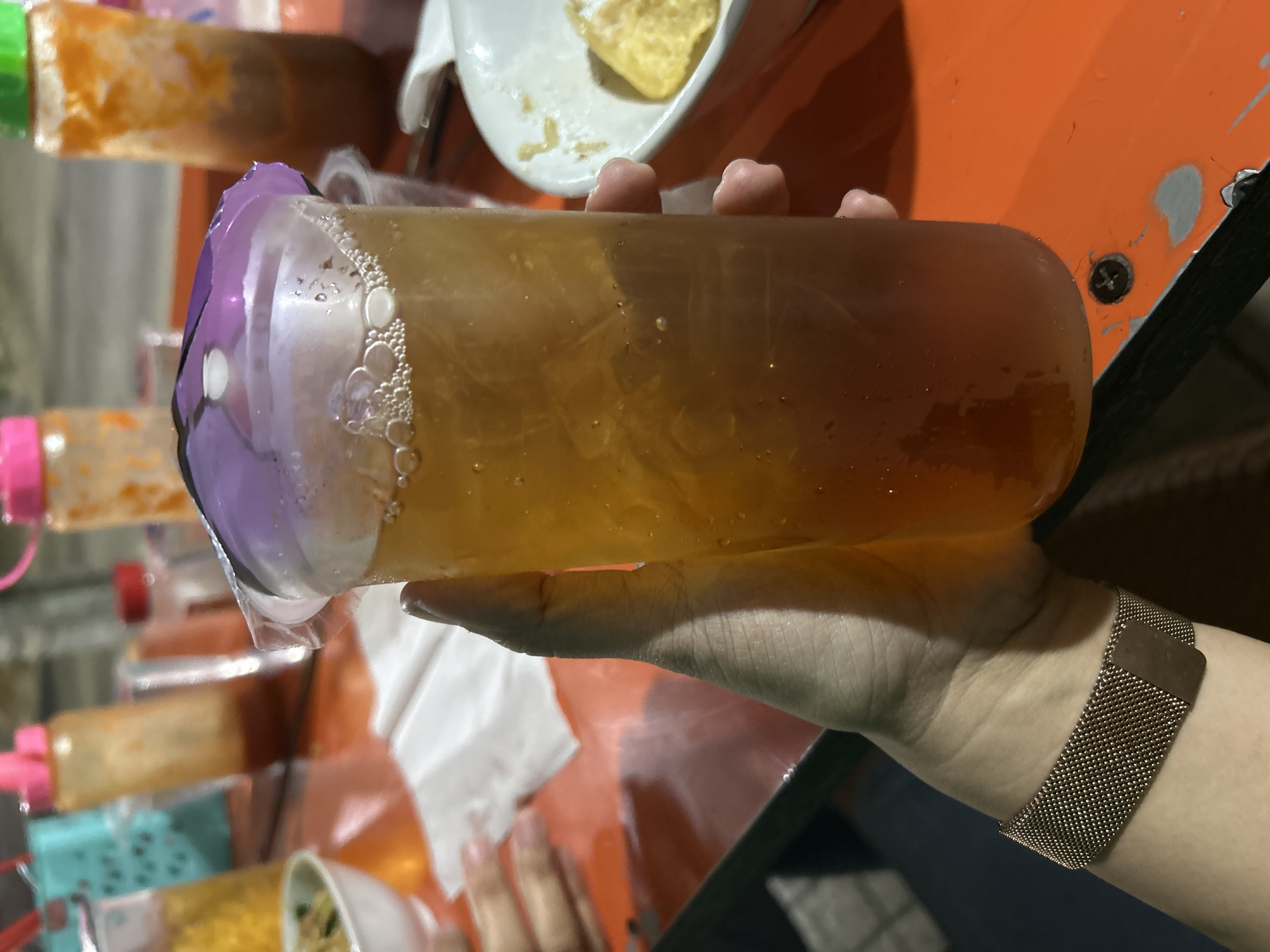 A person's hand is holding a clear plastic cup filled with iced tea. The cup has a purple plastic lid with a plastic seal. The tea is amber in color with ice cubes and small bubbles on the surface. The background shows an orange table with various items, including condiments in bottles, a white napkin, a bowl of food, and a wrist with a rose gold mesh watch band. The scene appears to be at an outdoor food stall or restaurant, possibly in Depok, Indonesia, given the context provided, implying an informal dining setting.