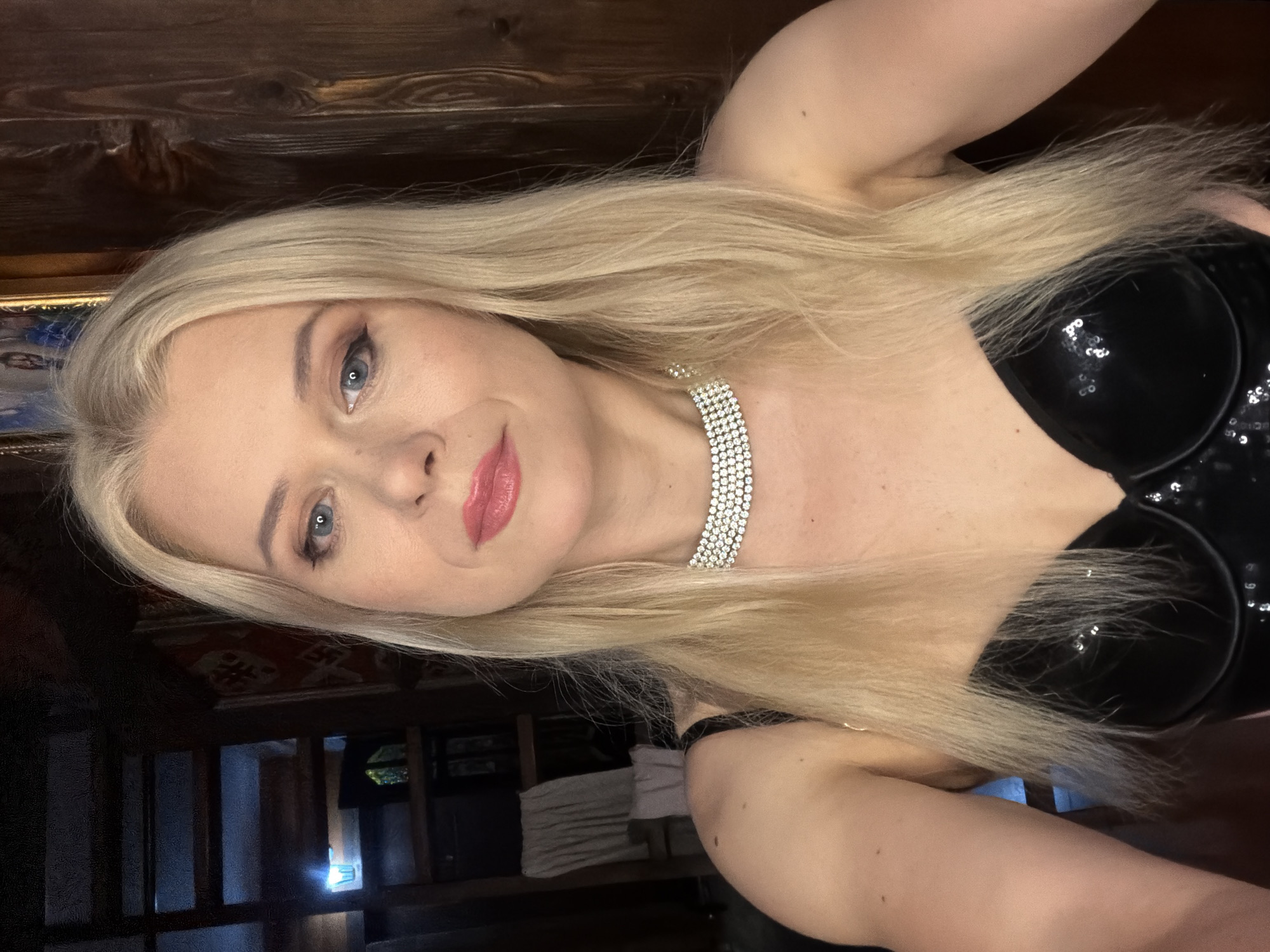 The image is a portrait of a blonde woman with blue eyes and pink lipstick, looking at the camera. She is wearing a black sequined top and a diamond-studded choker necklace. Her hair is long and blonde, and it falls over her shoulders. The background is a wooden wall and some shelves with objects on them, suggesting an indoor setting. The lighting appears to be artificial, creating a warm glow. The overall mood of the image is elegant and glamorous. No text is visible in the image. The location context is Sochi, Russia.