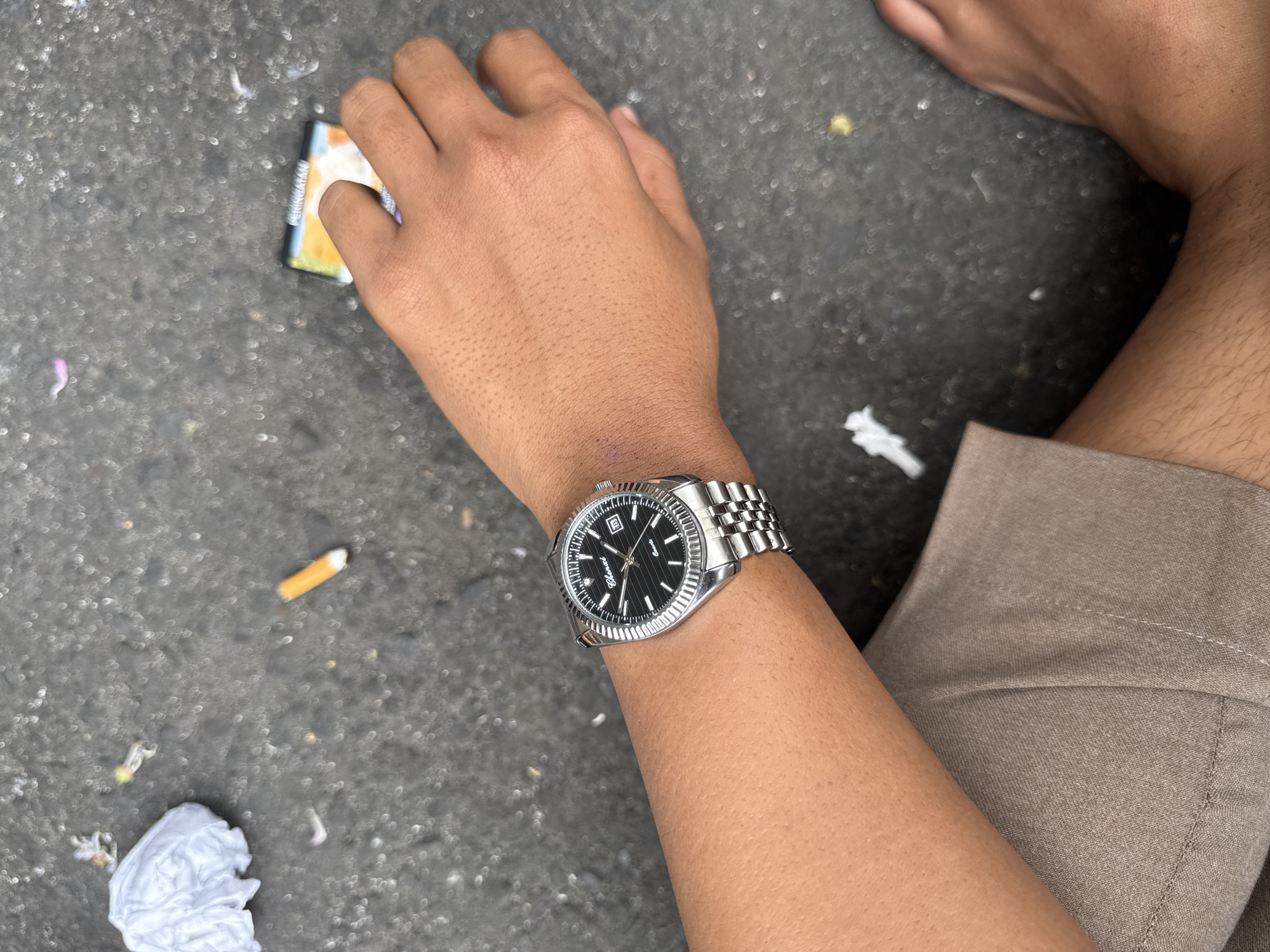 The image depicts a close-up, overhead view of a person's left arm and hand, adorned with a silver wristwatch, resting on a grey asphalt surface. The wrist shows a slight redness, possibly from the watch strap. The hand is relaxed with fingers slightly curled. Scattered across the asphalt are various debris, including crumpled white paper, cigarette butts, and what appears to be an open pack of cigarettes. The lighting suggests an outdoor environment, possibly daytime. The person is wearing beige-colored shorts, partially visible at the bottom of the frame. Another person's bare foot is visible in the lower right corner, with visible leg hair. The context of Kuta Selatan, Indonesia, implies an urban or semi-urban outdoor setting, consistent with the presence of street debris.