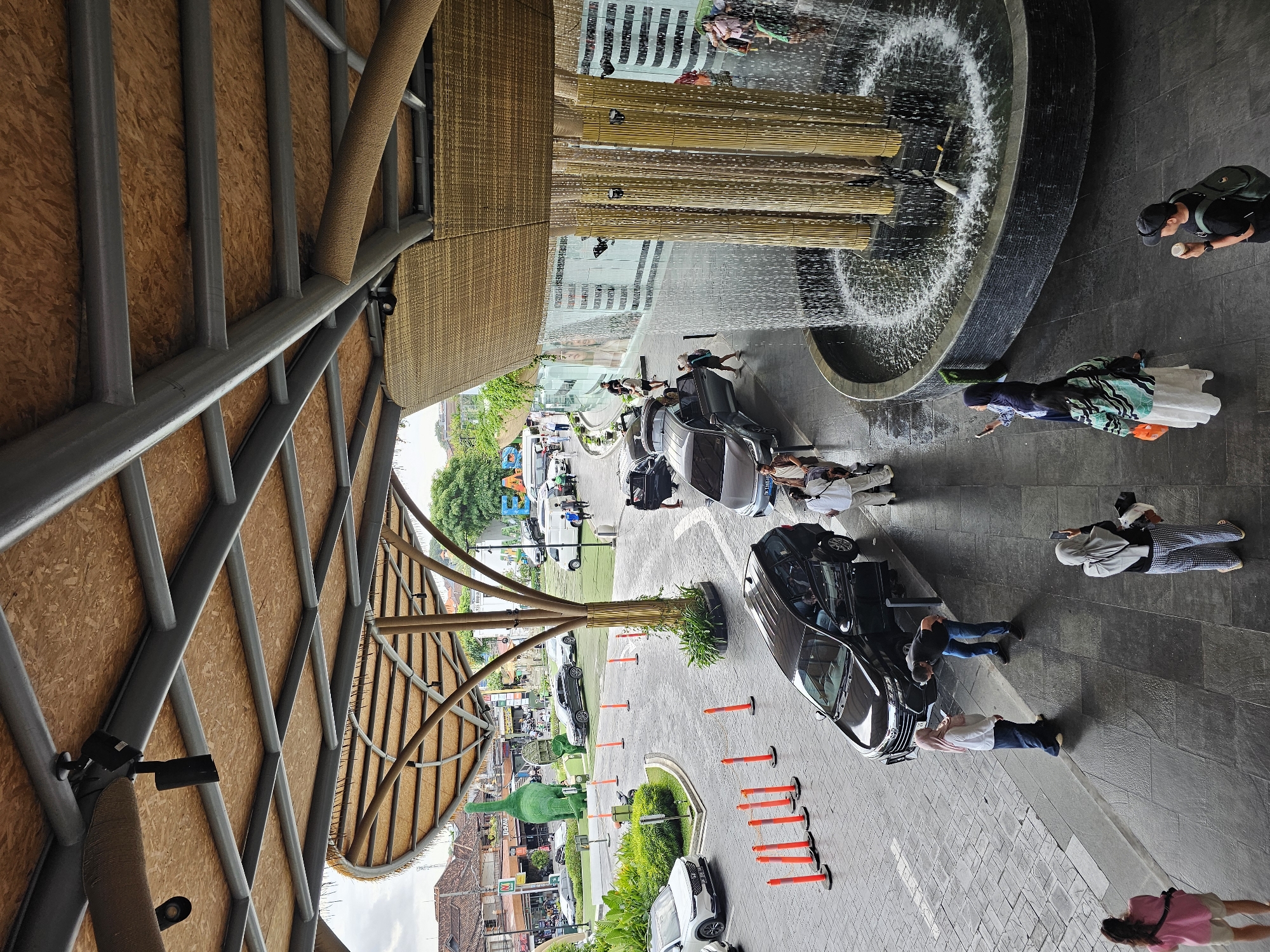 The image captures the entrance area of the Liv D'nDenpasar shopping mall in Denpasar, Indonesia. A large, open-air canopy with a thatched roof and exposed wooden beams covers the main entryway. Underneath, a circular fountain with bamboo-like structures sprays water into a basin. Several people are visible in the scene, some interacting with each other, others taking photographs. A black SUV is parked with its doors open, and a few other cars are in the background, along with a large green dinosaur statue and various shops. The ground is paved with stone tiles. The overall impression is that of a bustling public space with a contemporary design incorporating natural elements.