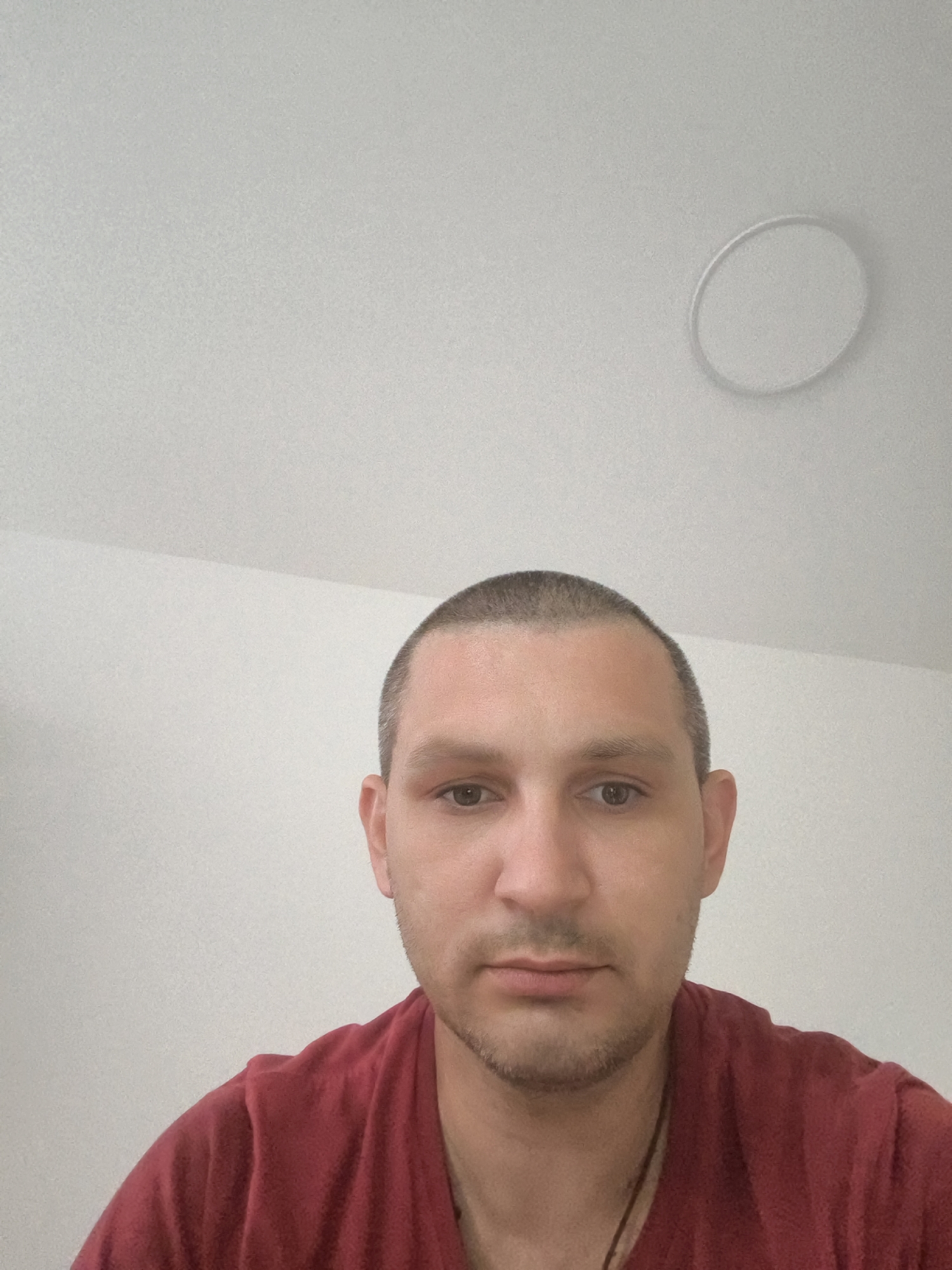 The image is a selfie taken indoors, likely in a room with a white ceiling and white walls. There is a circular fixture on the ceiling, possibly a light fixture or a smoke detector. The subject is a man with a buzzed haircut, light skin, and brown eyes. He is wearing a dark red t-shirt. He is looking directly at the camera. The overall impression is a casual, personal photograph. The image does not contain any information that would allow for the determination of a specific location within the city of Wolgast, Germany.