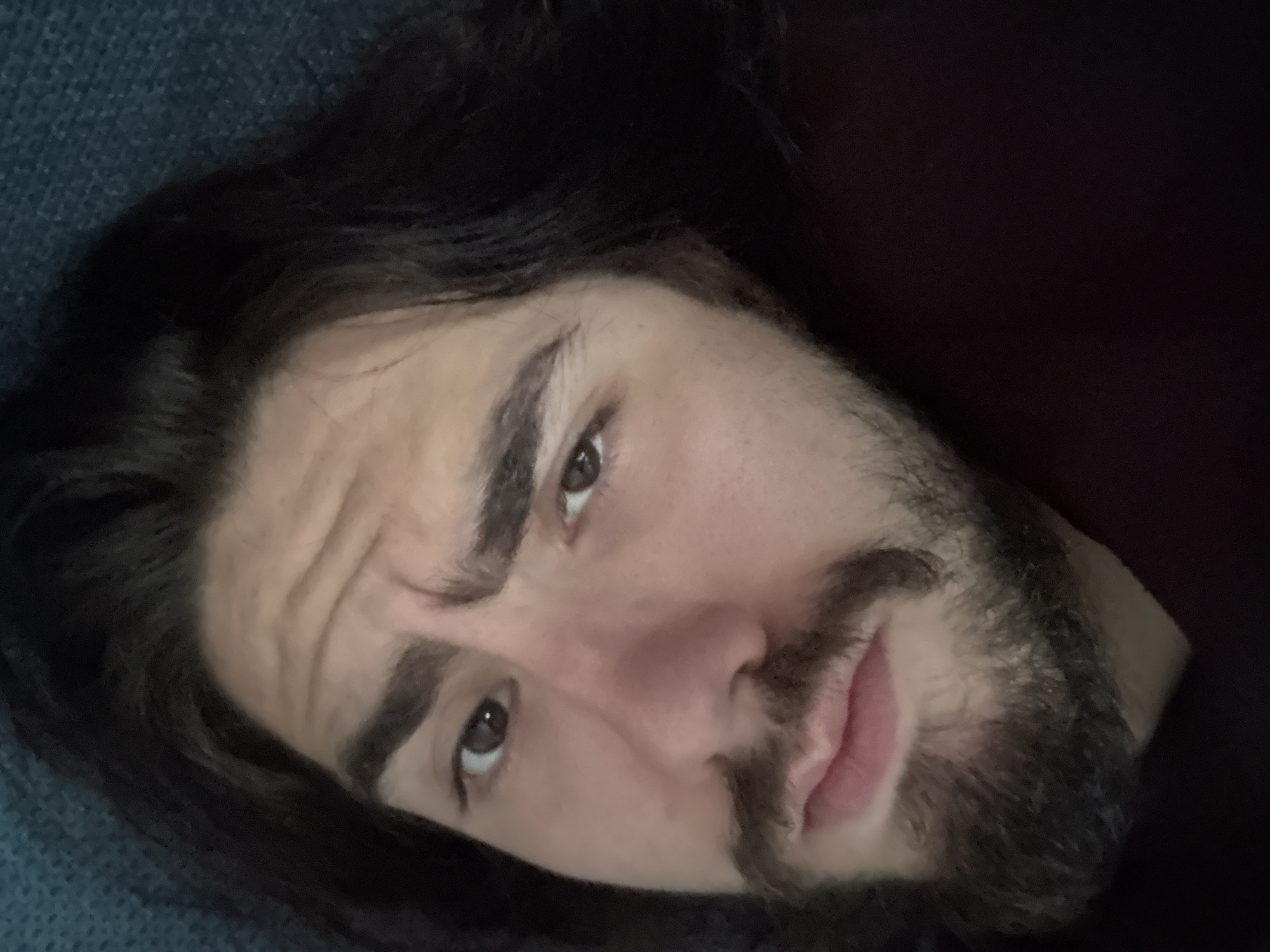 A close-up, overhead shot shows a young man with long, dark hair and a beard. He has dark brown eyes, a prominent nose, and thick eyebrows. His left eyebrow is furrowed, and his lips are slightly parted. The man is lying down, with his head tilted slightly to the right. The background is a dark, textured surface, possibly a couch or bed. The lighting is soft and diffused, casting subtle shadows on his face.