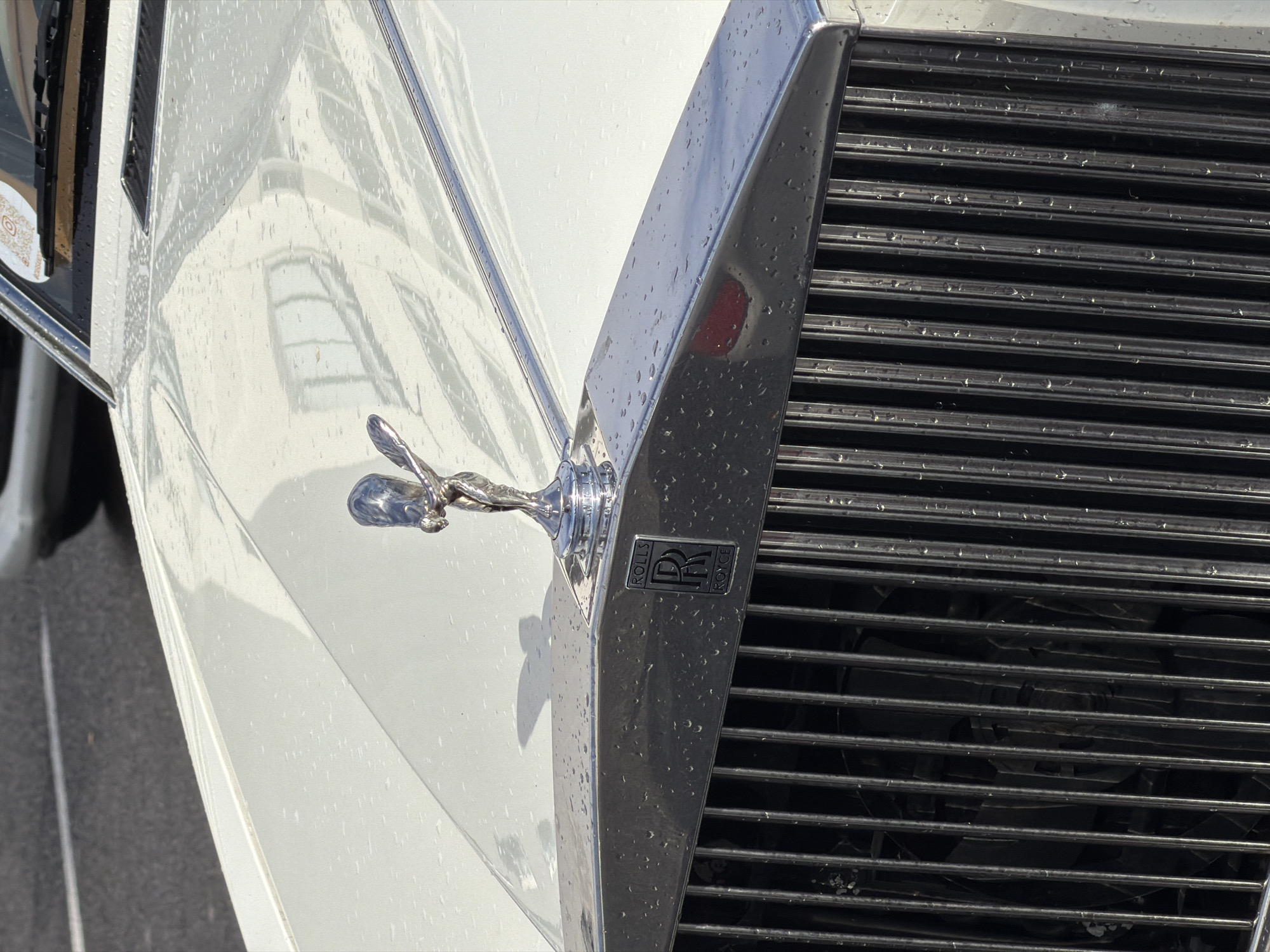 The image shows the front of a white Rolls Royce car. The iconic Spirit of Ecstasy hood ornament is visible atop the grill. The setting is indicated by the reflection of the Cape Town International Convention Centre in the car's paint, placing the location in Cape Town, South Africa. The vehicle has water droplets, suggesting it has either been raining or the car was recently washed.