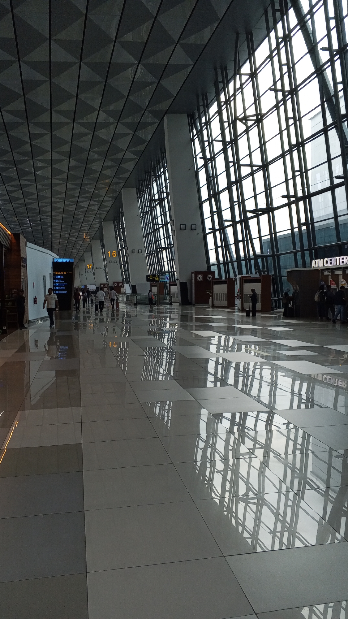 The image shows a long, wide hallway in an airport terminal in Rawa Burung, Indonesia. The hallway is lit by natural light from a large, floor-to-ceiling window on the right side of the image, which is covered by a grid of metal framing. The ceiling is a dark, geometric pattern of triangles. The floor is made of large, reflective tiles that create strong reflections of the window and ceiling structure. Several people are walking down the hallway in both directions. Some are walking alone, while others are in small groups. There are signs and displays visible along the walls, including a sign for "ATM CENTER". The overall impression is one of a modern, spacious, and busy transportation hub.