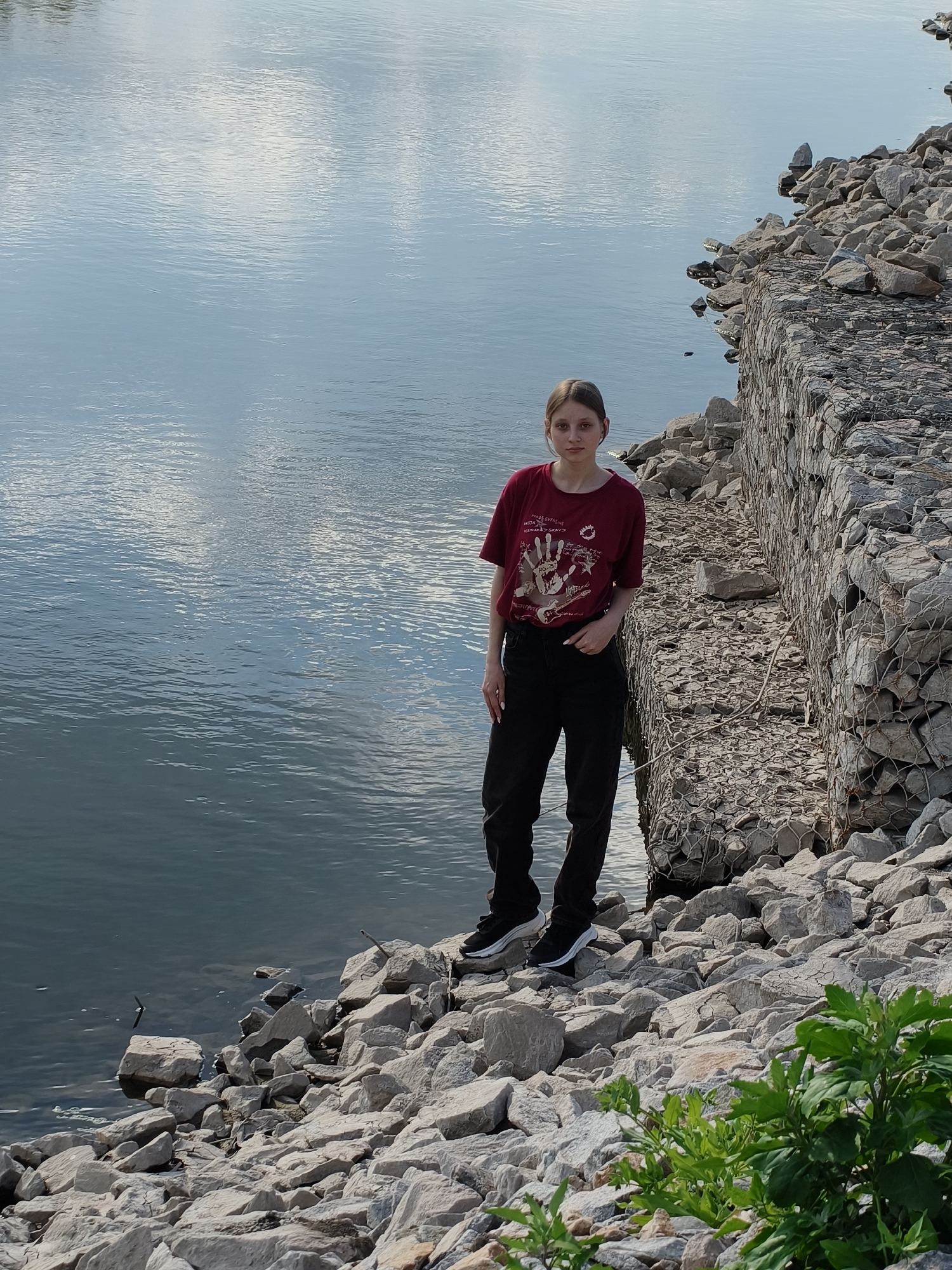 A full shot depicts a young woman standing on a rocky embankment next to a body of water. She is facing the camera with a neutral expression. She wears a dark red t-shirt with a design on it, dark pants, and black and white sneakers.
Behind and to her right is a rock wall reinforced with wire mesh. The water is a blue-gray color, reflecting the sky, which appears to have some cloud cover.
In the lower right corner is some green foliage. The embankment is made of various-sized gray rocks.
The location is the city of Kryvyi Rih, Ukraine.