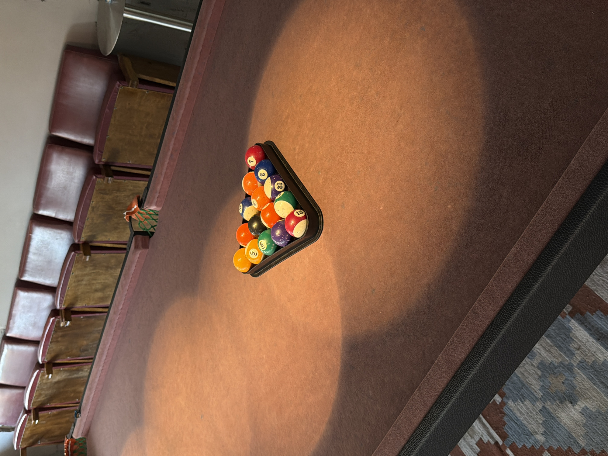 A billiard rack of balls is arranged on a brown felt table. Behind the table, a row of red theater seats is visible. The scene appears to be indoors, possibly in a private room or a recreational area. There are no people present in the image. The lighting suggests it might be daytime, with some natural light filtering in, casting shadows on the table.