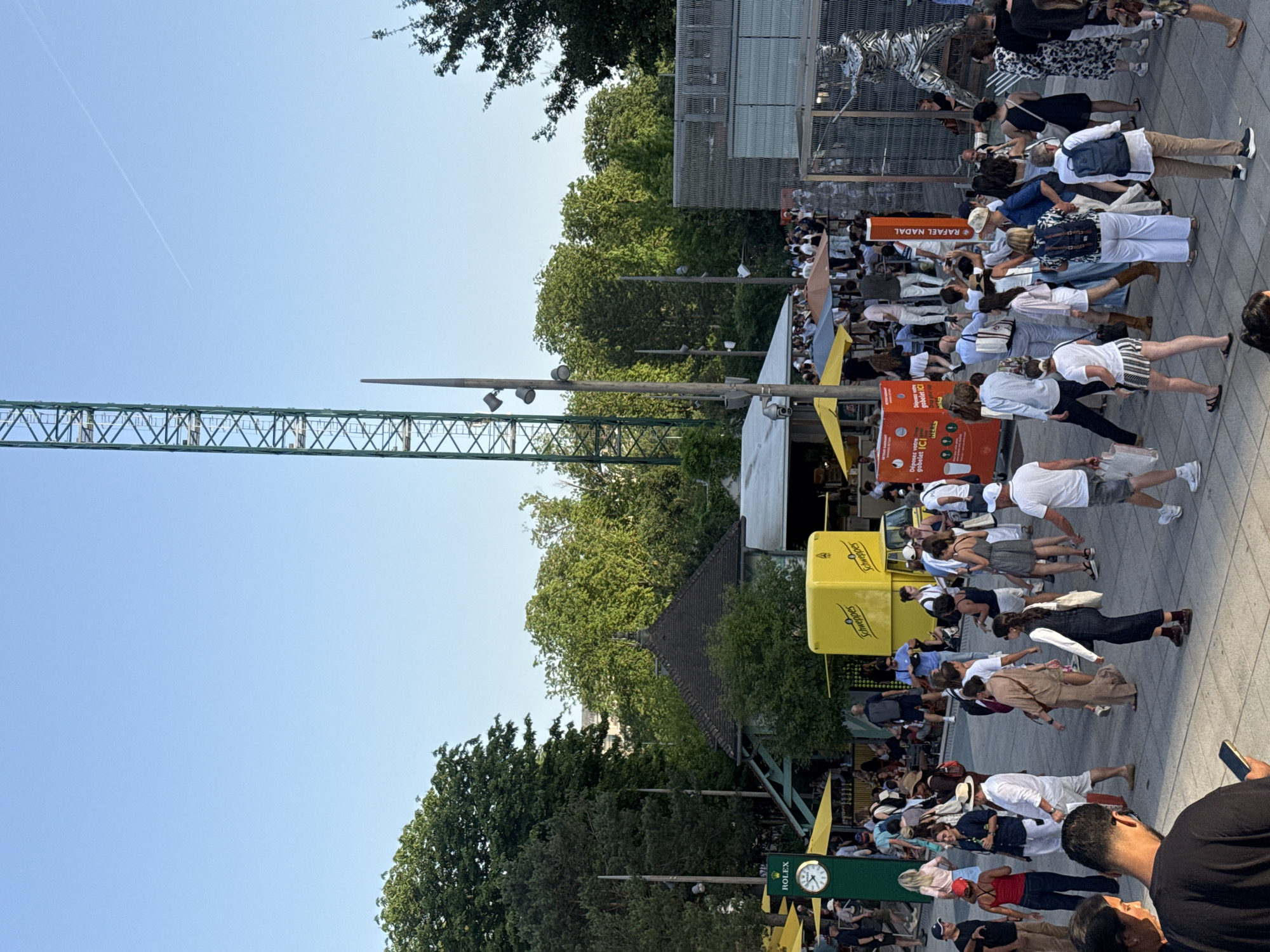 The image depicts a crowded outdoor scene at what appears to be a public event in Paris, France. A large group of people is walking along a paved area. Advertising banners for "Rafael Nadal," "Rolex," and "Schweppes" can be seen among the crowd, which suggest a sporting or tennis-related event, possibly the French Open at Roland Garros. A green crane structure stands tall on one side, partially obscured by trees. The weather appears to be sunny.