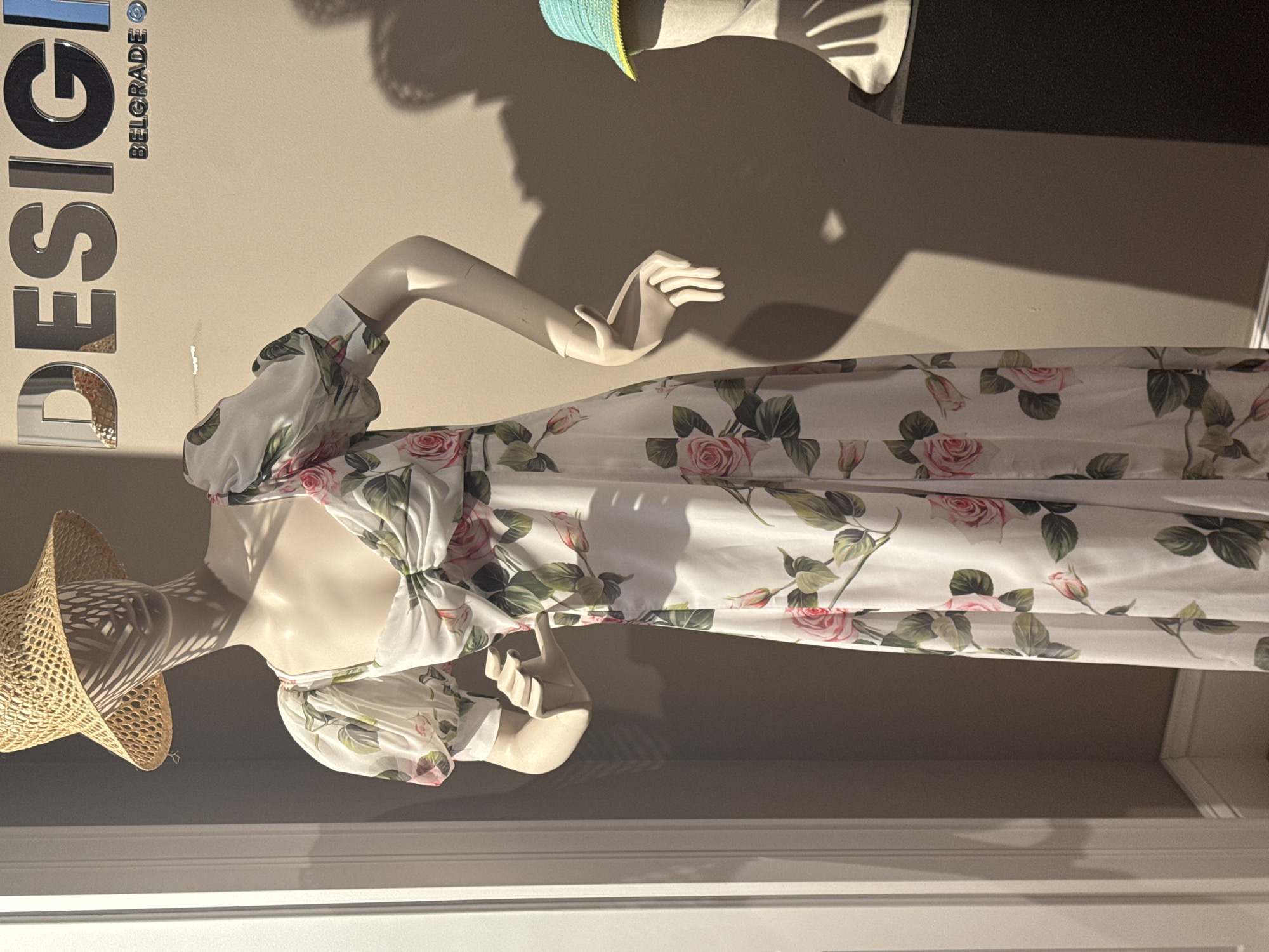 This media file showcases a fashion display in what appears to be a store or exhibition in Belgrade, Serbia. The central subject is a mannequin adorned with a flowing white dress featuring a delicate floral pattern of pink roses and green leaves. The dress has voluminous sleeves gathered at the shoulders and cuffs, and a cinched waist with a large bow. The mannequin is also wearing a wide-brimmed straw hat that casts a patterned shadow on its face.
To the left of the mannequin, large, metallic letters spell out "DESIGN" vertically, with the word "BELGRADE" written horizontally beneath it. The setting is a well-lit interior with a neutral-colored wall, suggesting a retail or gallery environment. Shadows from the mannequin and possibly other display elements are cast across the wall, indicating artificial lighting, likely from above. The overall impression is one of a sophisticated and curated presentation of clothing and design.