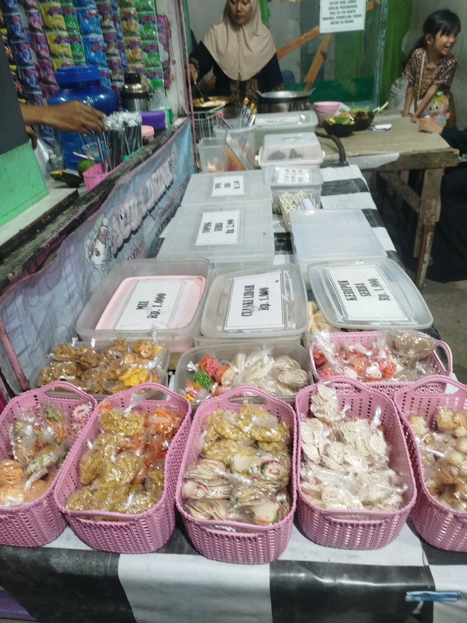 In the city of Burneh, Indonesia, a market stall displays a variety of snacks. The stall is adorned with a banner featuring cartoon characters. Behind the counter, a woman in a beige hijab and a dark patterned dress is cooking at a stove. To her right, a young girl in a similar patterned dress with a stuffed toy slung over her shoulder looks to her left, smiling.
In the foreground, several pink woven baskets are filled with individually packaged snacks. These snacks appear to be a mix of crunchy items, some golden brown and others white. Some plastic containers on the counter also hold individually packaged snacks with labels indicating prices in Indonesian Rupiah. One label reads "MIE Rp. 1.000". Other visible labels include "KERUPUK KEPALA" and "KERUPUK UDANG". Behind the counter, shelves are stocked with various bagged snacks. A large blue water dispenser is visible to the left. The overall environment suggests a busy, open-air market setting.