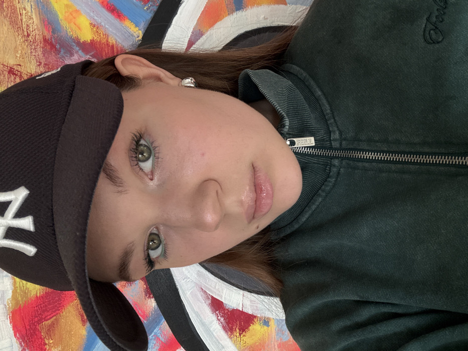 A close-up portrait of a young individual, likely female, is captured in Vozdovac Urban Municipality, Serbia. The subject possesses fair skin, green eyes, and light brown hair partially visible beneath a dark baseball cap with a prominent white "NY" logo. She maintains direct eye contact with the camera, displaying a neutral facial expression with full, glossy lips and a small mole situated below her lower lip to the left. She is dressed in a dark green, collared zip-up jacket, which appears somewhat faded. The jacket features a silver-toned zipper with a pull tab embossed with "PULL", and faint embroidered text, possibly "Foots", is visible on the lower right front. A small, reflective silver-toned hoop earring adorns her visible right earlobe. The background is an abstract painting or mural composed of bold brushstrokes and shapes in bright colors including red, yellow, and blue, contrasted with black and white elements, notably a large black arc with a white outline on the left.