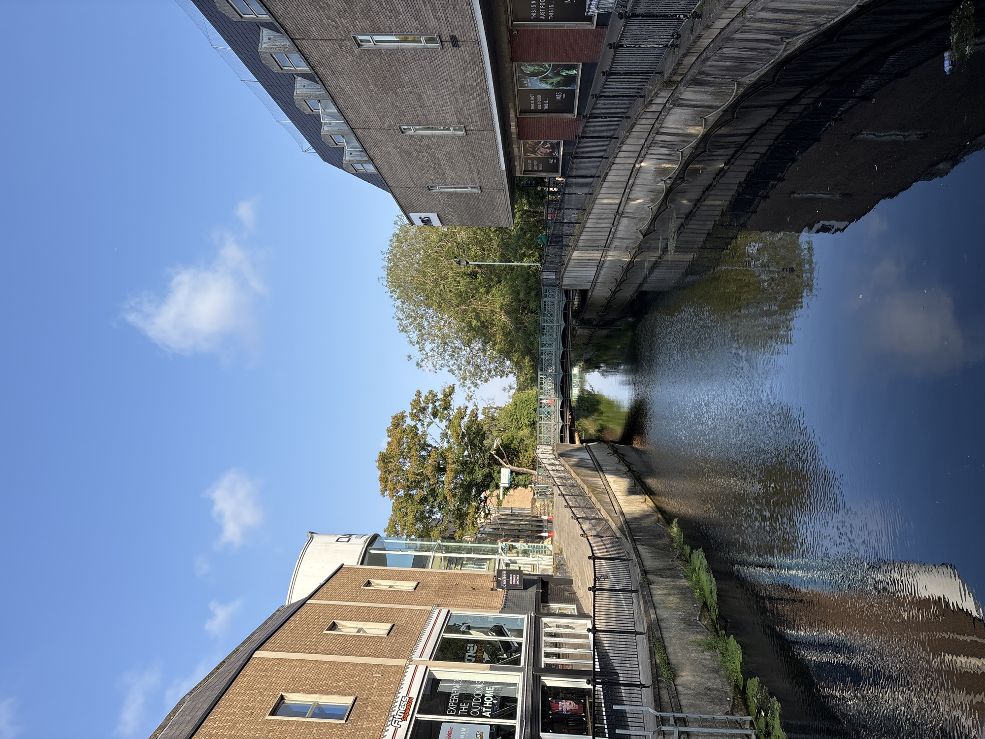 This is a vertical, eye-level shot of the River Chelmer in Chelmsford, United Kingdom, on a clear, sunny day. The river flows from the background towards the foreground, reflecting the blue sky with scattered white clouds and the surrounding buildings. On the left bank, a brick building with large glass windows, housing a "Fitness for Less" store, is visible. Further along, a modern white curved structure with the partial text "DA" is apparent. On the right bank, a brick building with a darker, sloped roof houses an "M&S Foodhall" store. A pedestrian bridge spans the river in the middle distance, with trees and more buildings visible beyond it. The riverbanks are lined with grey concrete walls and black metal railings. Some green algae is visible floating on the water's surface. The scene appears tranquil, with no people actively engaged in any discernible activities.