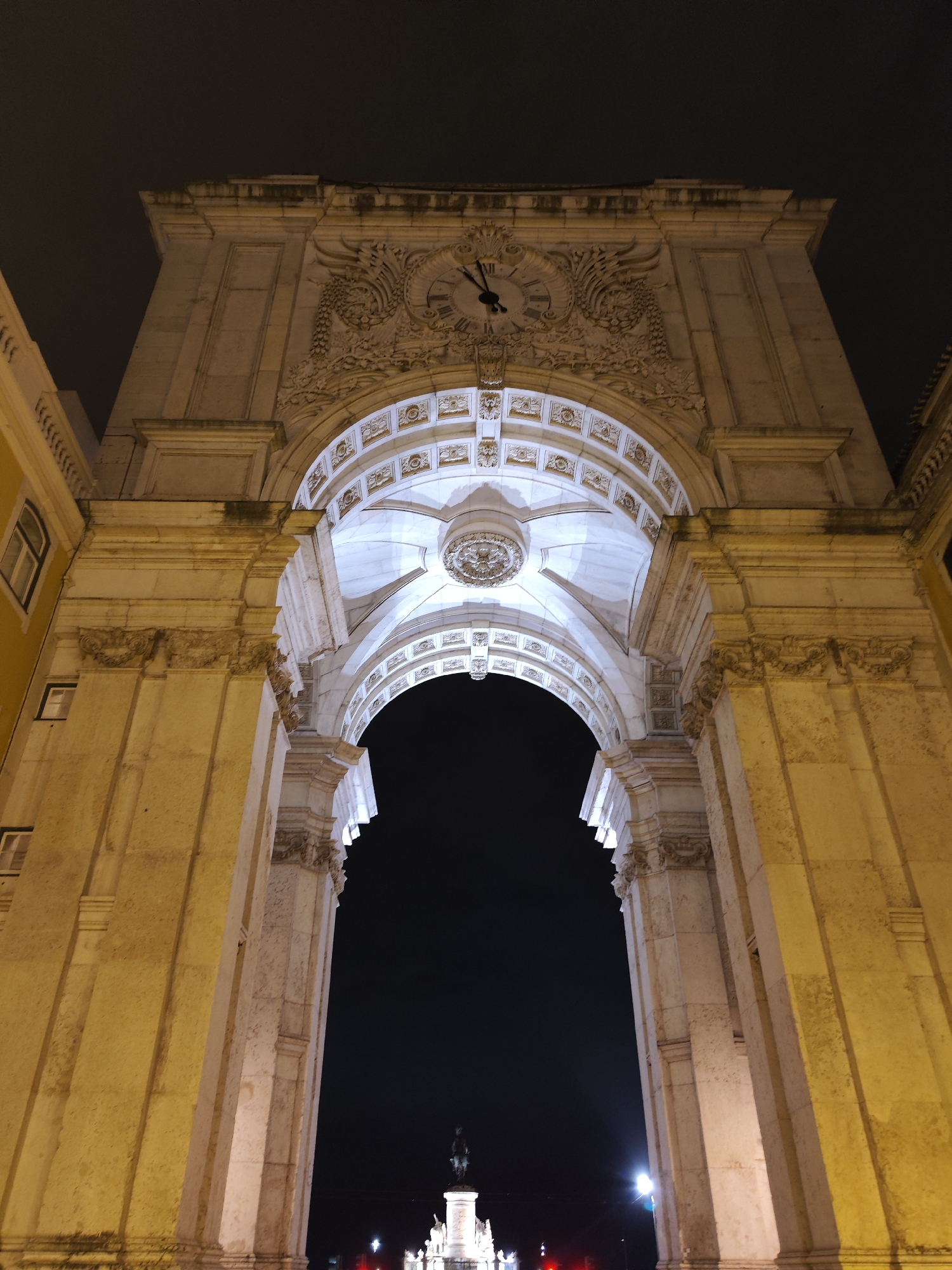 The image captures a majestic, ornate stone archway at night, viewed from a low-angle perspective looking upwards. The grand structure, characteristic of neoclassical or Baroque architecture, is intricately carved with decorative motifs, including scrolling foliage, columns, and cornices. A large clock face with Roman numerals (I through XII) is integrated into the frieze above the main arch, displaying the time as approximately 10:10 or 10:11.
The archway is dramatically illuminated by artificial lighting, casting a warm, golden glow on its exterior surfaces, highlighting the textures of the stone and the detailed carvings. The underside of the arch is lit with a cooler, white light, emphasizing its coffered ceiling and a central medallion. Through the dark void of the arch, a brightly lit equestrian statue on a pedestal is faintly visible in the distance, centered beneath the arch.
To the left, a portion of an adjacent building with several windows is visible, painted in a distinct yellow or ochre hue, also illuminated. The sky above is dark, indicating nighttime. The scene conveys a sense of historical grandeur and prominence, typical of a significant city landmark, which is known to be the Arco da Rua Augusta in Lisbon, Portugal, leading into Praça do Comércio.
