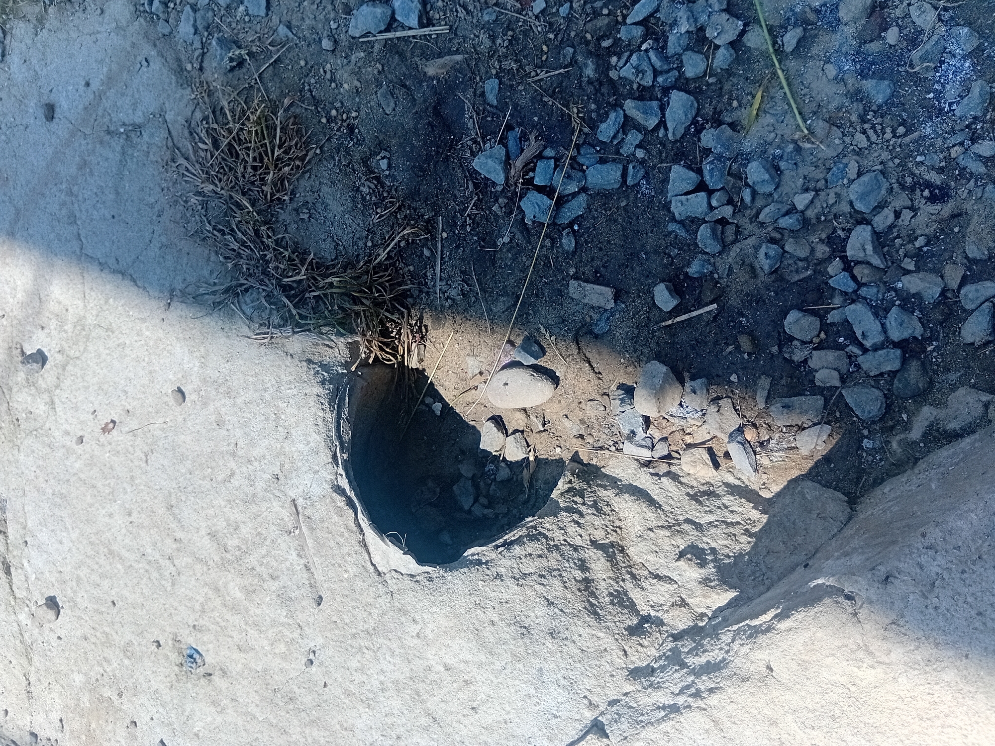 This media file shows a close-up, overhead view of a section of concrete with a hole in it, next to a gravelly area. The concrete is rough and appears to be a pathway or sidewalk. The hole is circular and dark, with rocks and dirt visible inside. To the left of the hole, a clump of dry, brown grass is growing out of the concrete. The gravelly area to the right is composed of various sized grey and blue stones, along with dirt and dried plant matter. A single, smooth, light-colored stone rests on the edge of the hole. Sunlight casts a strong shadow across the scene from the left. The overall impression is of an outdoor, possibly rural or undeveloped area. There is no visible text.