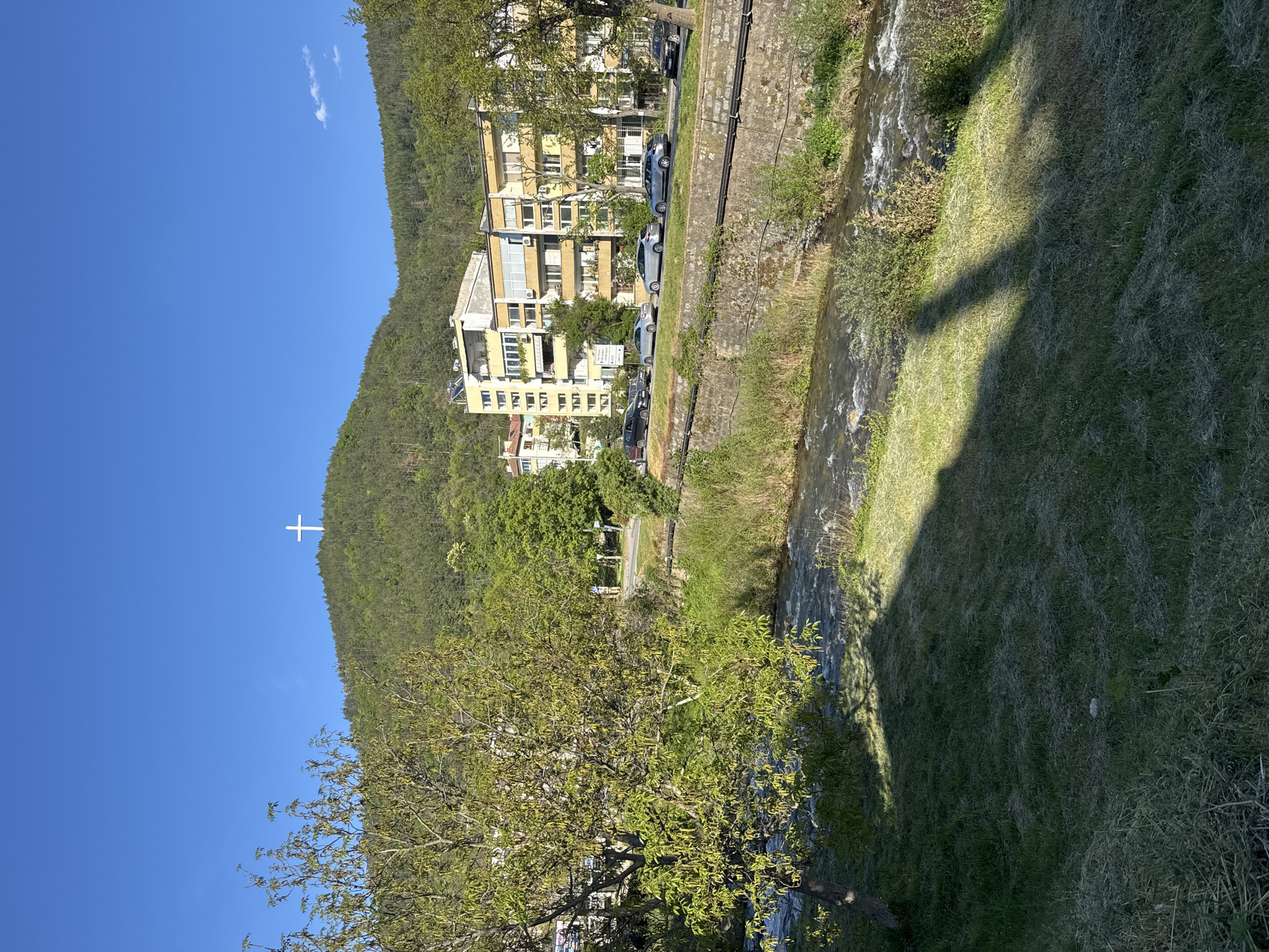 The image depicts an outdoor scene in Blagoevgrad, Bulgaria. The main features are a river running along the side of the frame, with buildings and cars lined up along its bank. On the opposite side of the frame, there is a grassy area with a large shadow cast on it. In the background, a hill covered in greenery rises, with a large white cross visible near its summit. The sky is a clear blue. A tree is partially visible in the foreground.