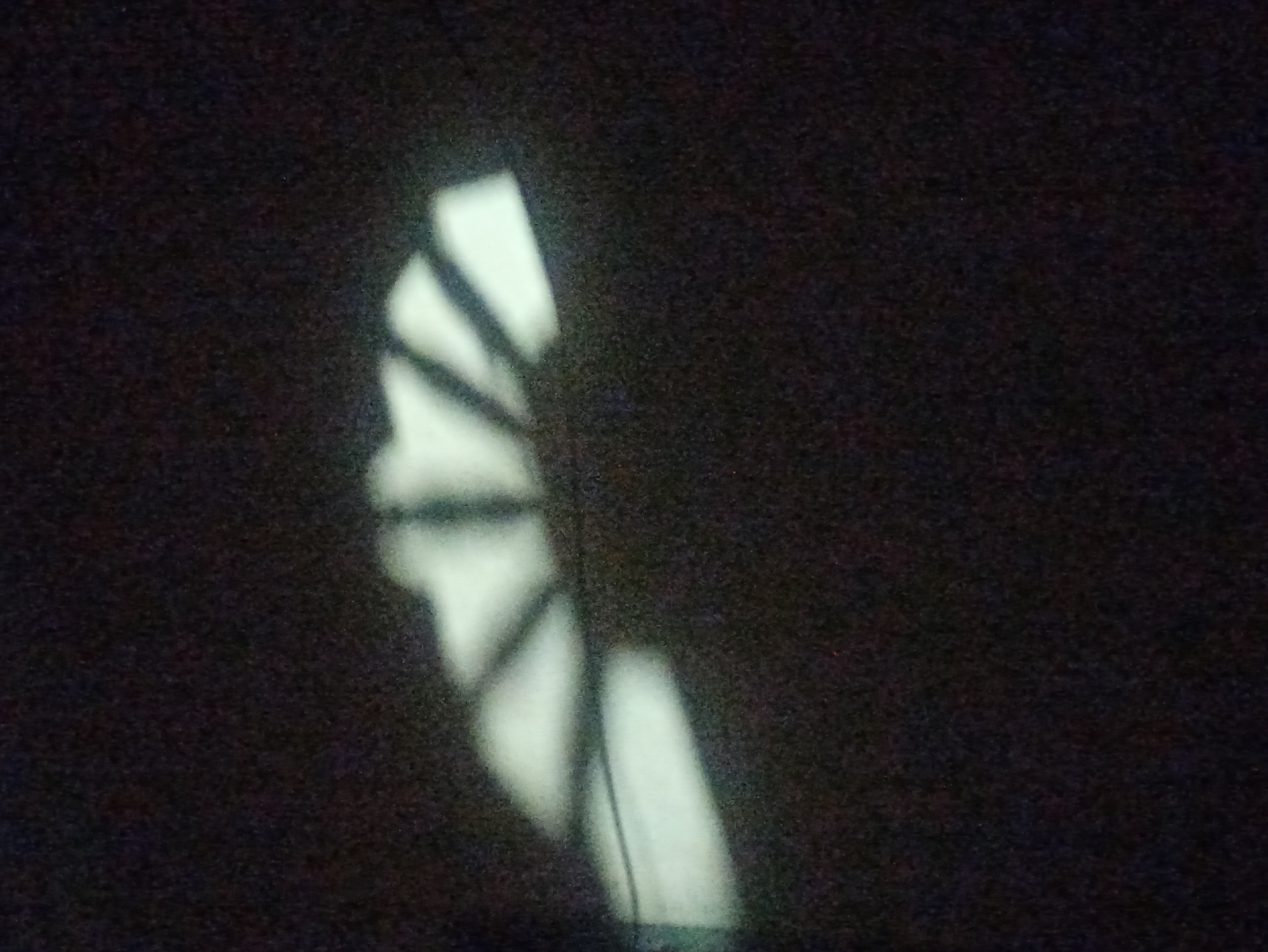 The image shows a dark background with a light-colored shadow cast on it. The shadow appears to be from a fan or a radiator, with several radiating lines forming a semi-circular shape. The texture of the background is rough and granular. There are no people or discernible objects other than the shadow. The scene is indoors, with the light source likely coming from a window or a lamp that is projecting the shadow onto a surface like a wall or floor. The image does not provide any information that would allow for the identification of its location in Tuba, Indonesia.