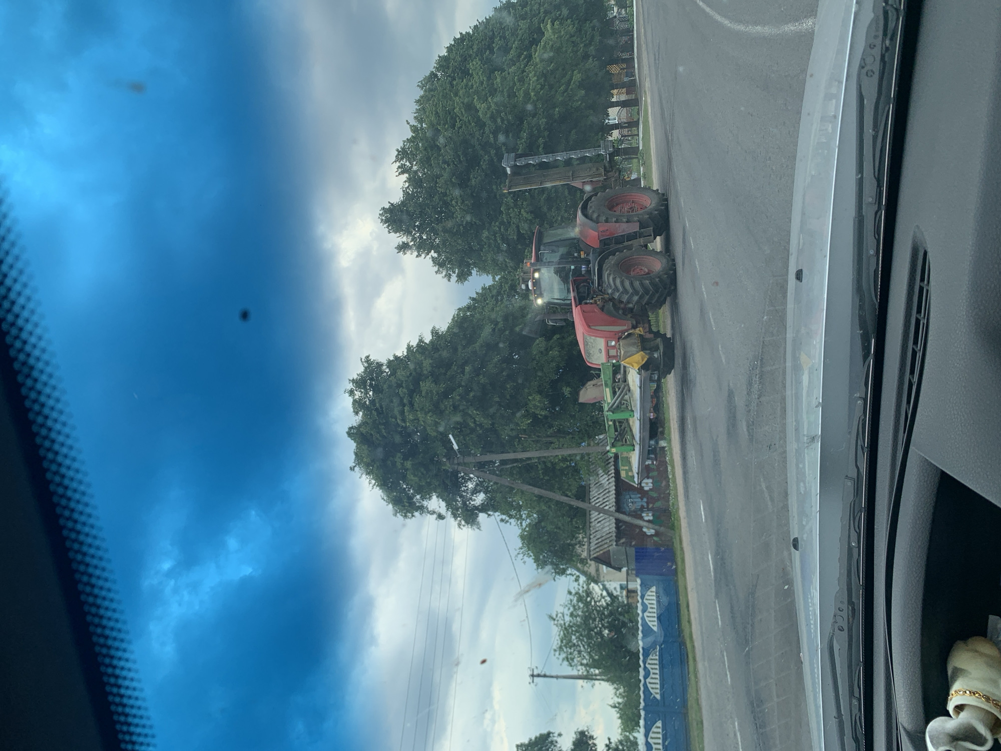 The image shows a red tractor with a harvesting attachment on a road in Слабада-Кучынскі сельскі Савет, Belarus. The tractor is moving on the right side of the road, with trees lining the background. The sky is cloudy, and the overall lighting suggests it is daytime. A blue fence with a white decorative pattern runs along the side of the road. A small building with a gray roof is visible behind the fence, and some graffiti is present on the building's wall. The foreground shows the dashboard and windshield of a vehicle, indicating the image was taken from inside a car. The windshield is slightly dirty.