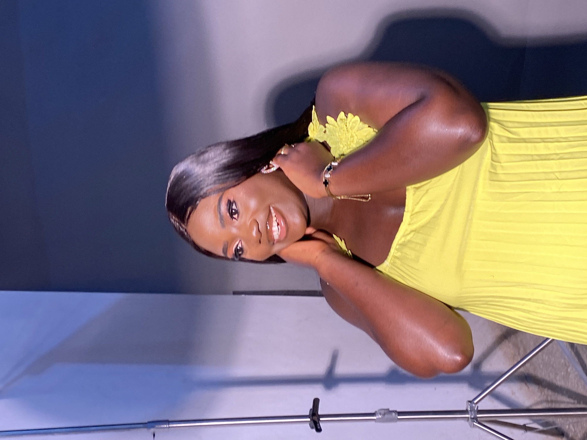A medium shot, captured from a slightly low angle, shows a Black woman standing in what appears to be a studio setting. She is wearing a bright yellow dress with a pleated skirt and floral detailing on the shoulder straps. Her hands are raised to her neck, and she is looking off to the right with a wide smile and her mouth open. She has long, dark hair, winged eyeliner, and bold eyebrows. On her left wrist, she wears a delicate gold bracelet with black accents. To the left of the frame, a metal tripod with a light stand is visible. The background is a gradient of light blue to dark blue, suggesting a studio backdrop. The lighting is focused on the woman, highlighting her features and the texture of her dress. There is no discernible location in Gura Top, Nigeria.