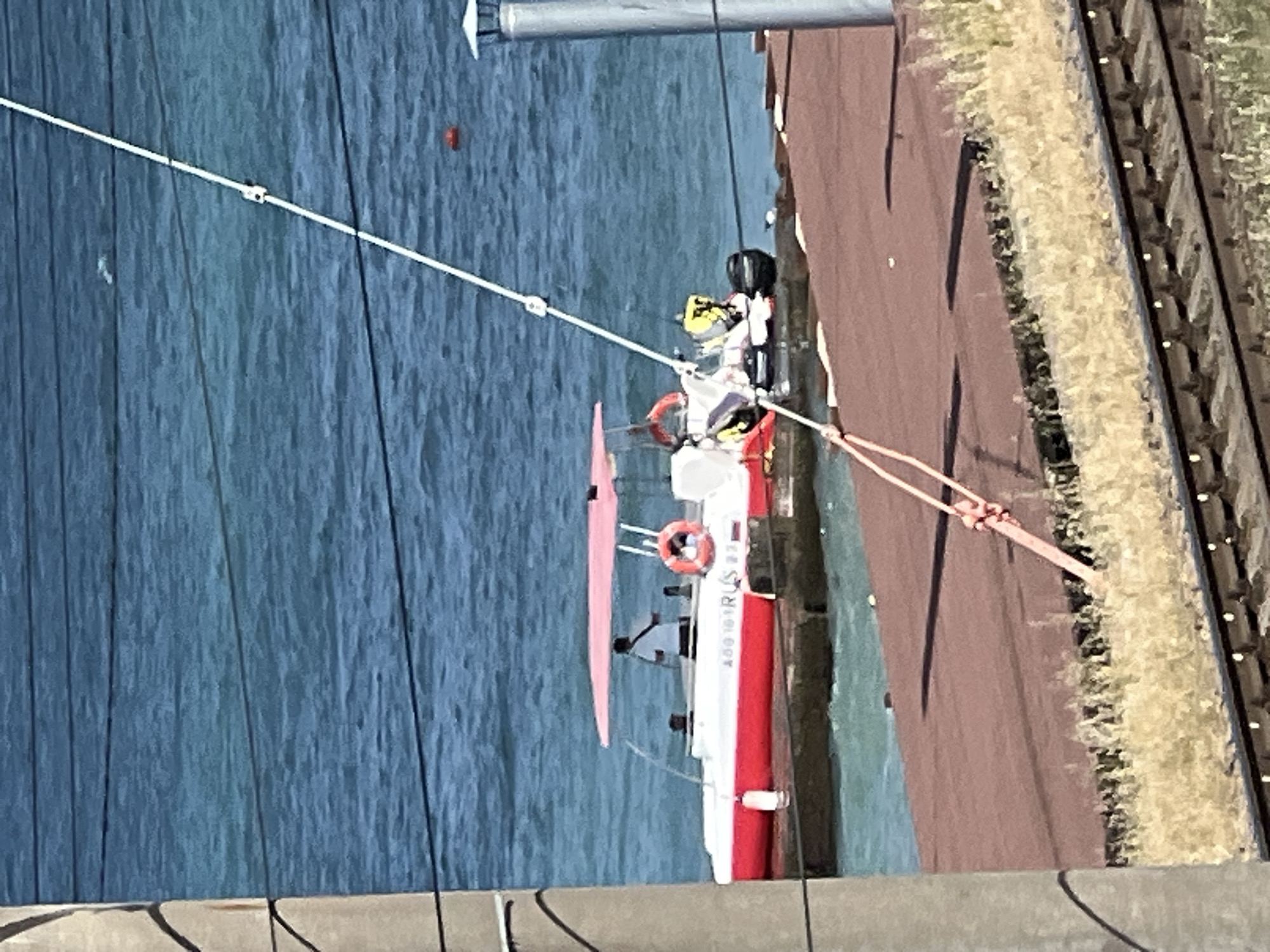 The image shows a red and white boat with a pink canopy, identified by the inscription "A801B1 RUS 22", docked at a wooden pier. A person wearing a white shirt stands on the boat. The boat is on a body of blue water, likely a sea or a large river. In the foreground, there are train tracks and dry grass. A tall metallic structure, possibly a lamppost or a part of a railway signal, is visible on the right. The scene is bathed in bright sunlight, casting shadows on the pier. The presence of train tracks suggests a location near a railway line. The inscription "RUS" and the overall aesthetic are consistent with a scene in Russia, possibly near a coastal area like Sochi, which is known for its proximity to the Black Sea and its railway infrastructure.
