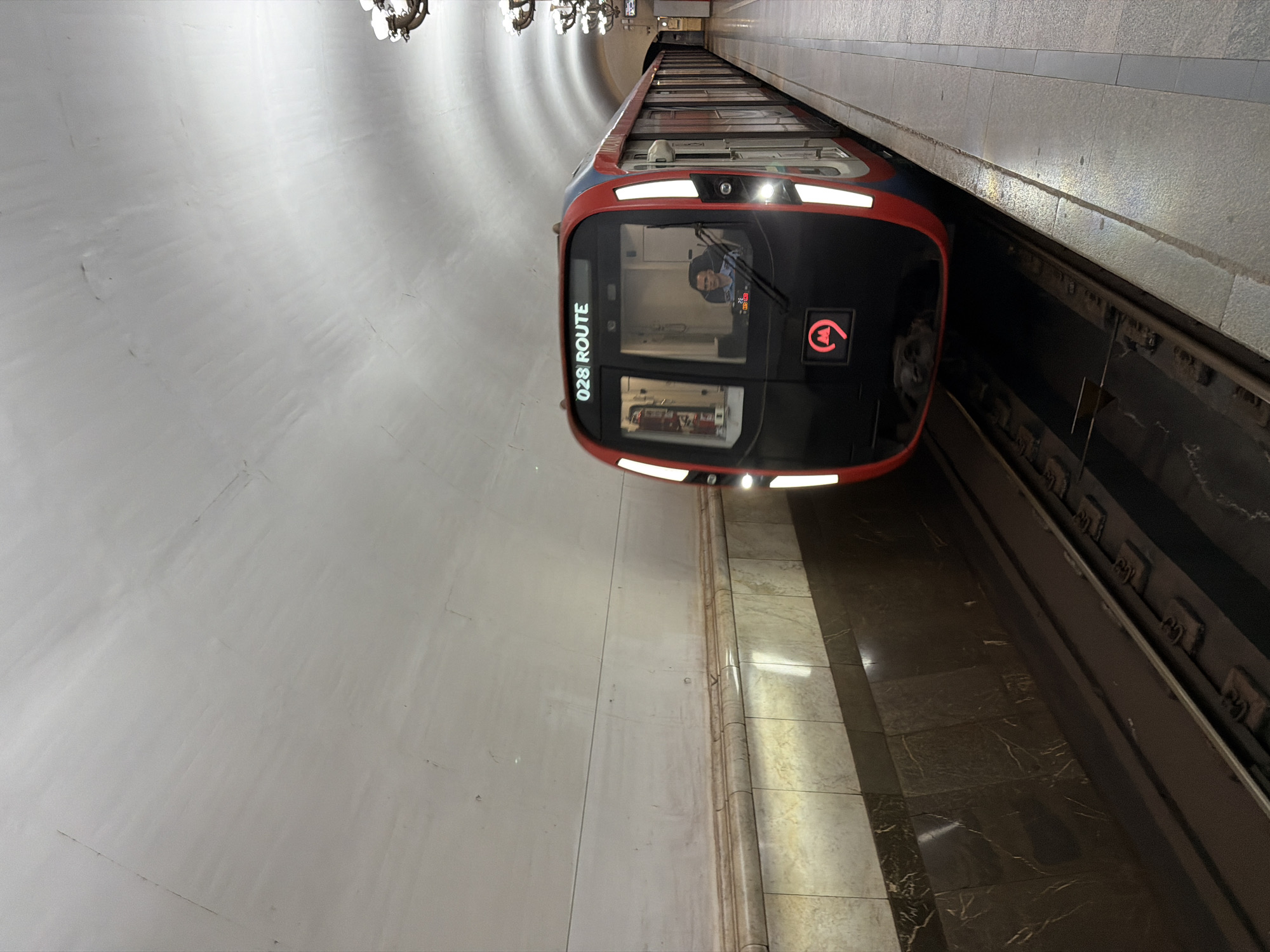 A low-angle, rotated, medium shot depicts the front of a red and black metro train with the words "028 ROUTE" on the windshield, likely in a station on the Moscow Metro in Russia. The train's headlights are illuminated, and a male driver is visible in the operator's compartment. The station has a curved, white ceiling with ornate chandeliers visible in the distance. A marble-like platform is along the right edge of the image, adjacent to the train tracks.