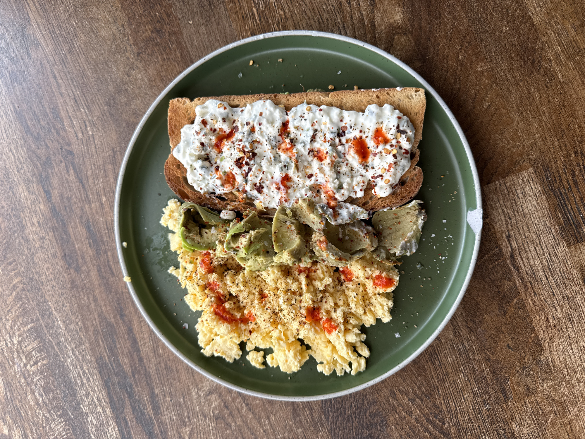 A top-down shot reveals a green plate on a dark wooden surface. The plate holds three distinct food items. First, a piece of toast is topped with a white creamy spread sprinkled with red pepper flakes and droplets of red sauce. Below the toast is a pile of scrambled eggs with red sauce and seasoning. To the side of the eggs sits sliced avocado. There is no evidence to indicate this photo was taken in Stotfold, United Kingdom.
