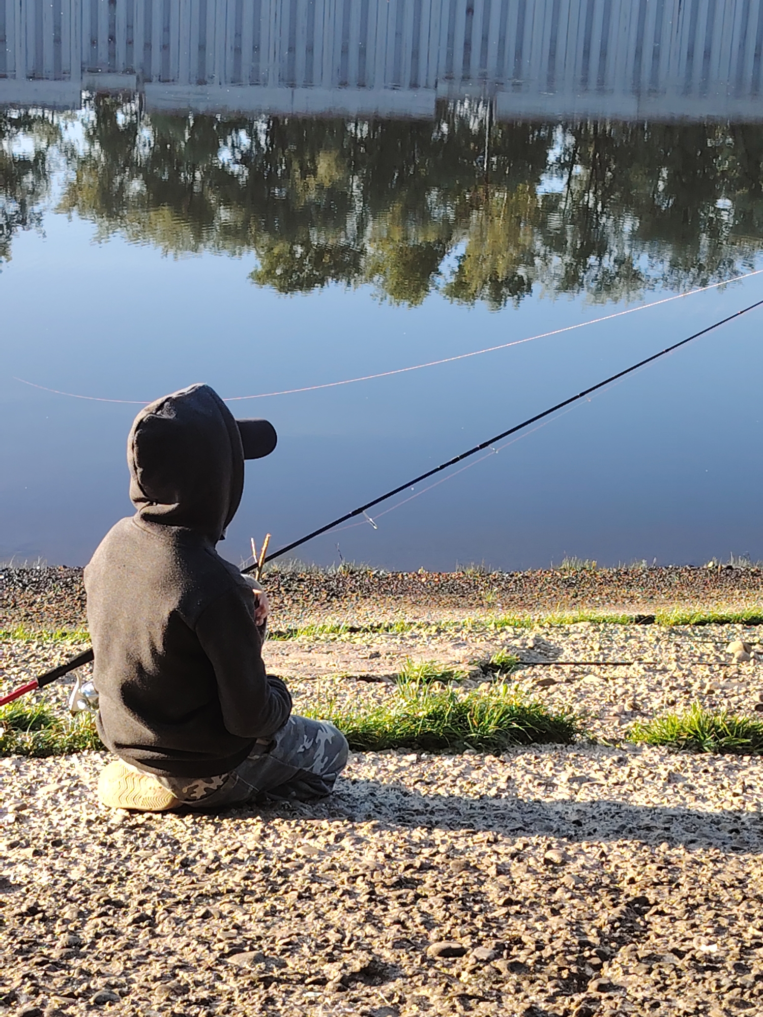 An individual wearing a dark hooded sweatshirt and camouflage pants sits on a gravelly shoreline, facing a calm body of water. A fishing rod rests in front of them, its line extending into the water. The reflection of a fence and trees is visible on the water's surface. The setting appears to be a natural area near a body of water within the city of Chaikovsky, Russia.