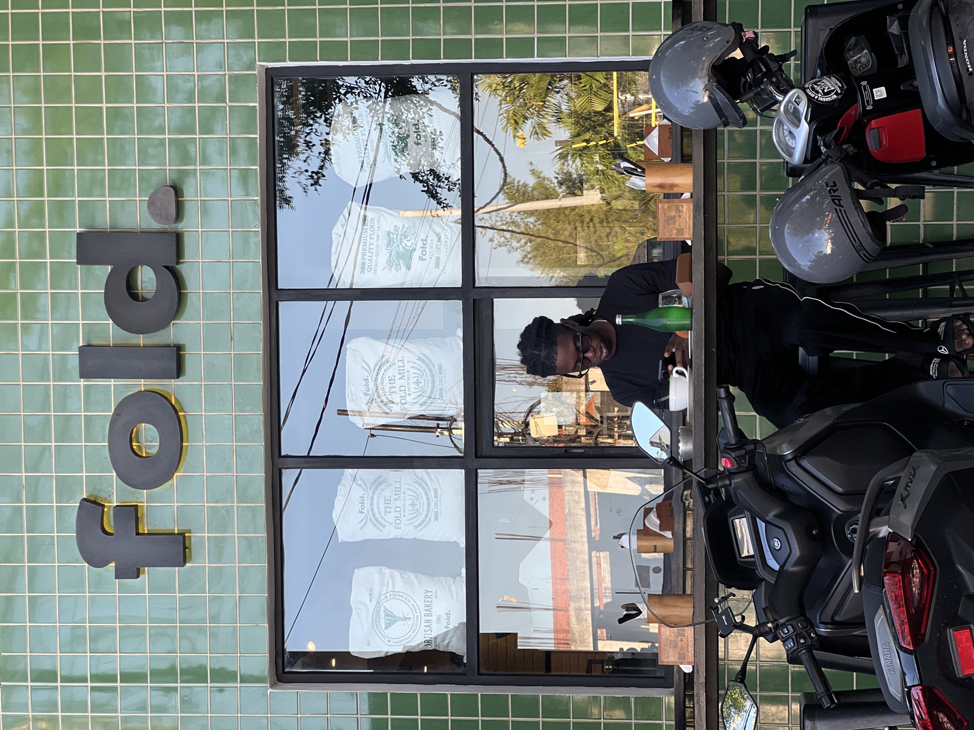 The image captures a scene at a "fold." bakery, as indicated by the sign on the green tiled wall. The bakery's window reflects the exterior environment, including utility lines, trees, and what appears to be the reflection of another building. Inside, a person is seated, pouring from a green bottle into a glass or cup. The window display features multiple bags of "fold." Premium Quality Flour from The Fold Mill, indicating they may produce or use their own flour. A parked Yamaha Xmax scooter and Honda scooter, both with helmets, are visible outside, suggesting the location is accessible by scooter. The branding on the flour bags includes the text "BALI, INDONESIA" placing the location in Bali, Indonesia. Based on the other landmarks and the information present in the image, this bakery is located in North Kuta.