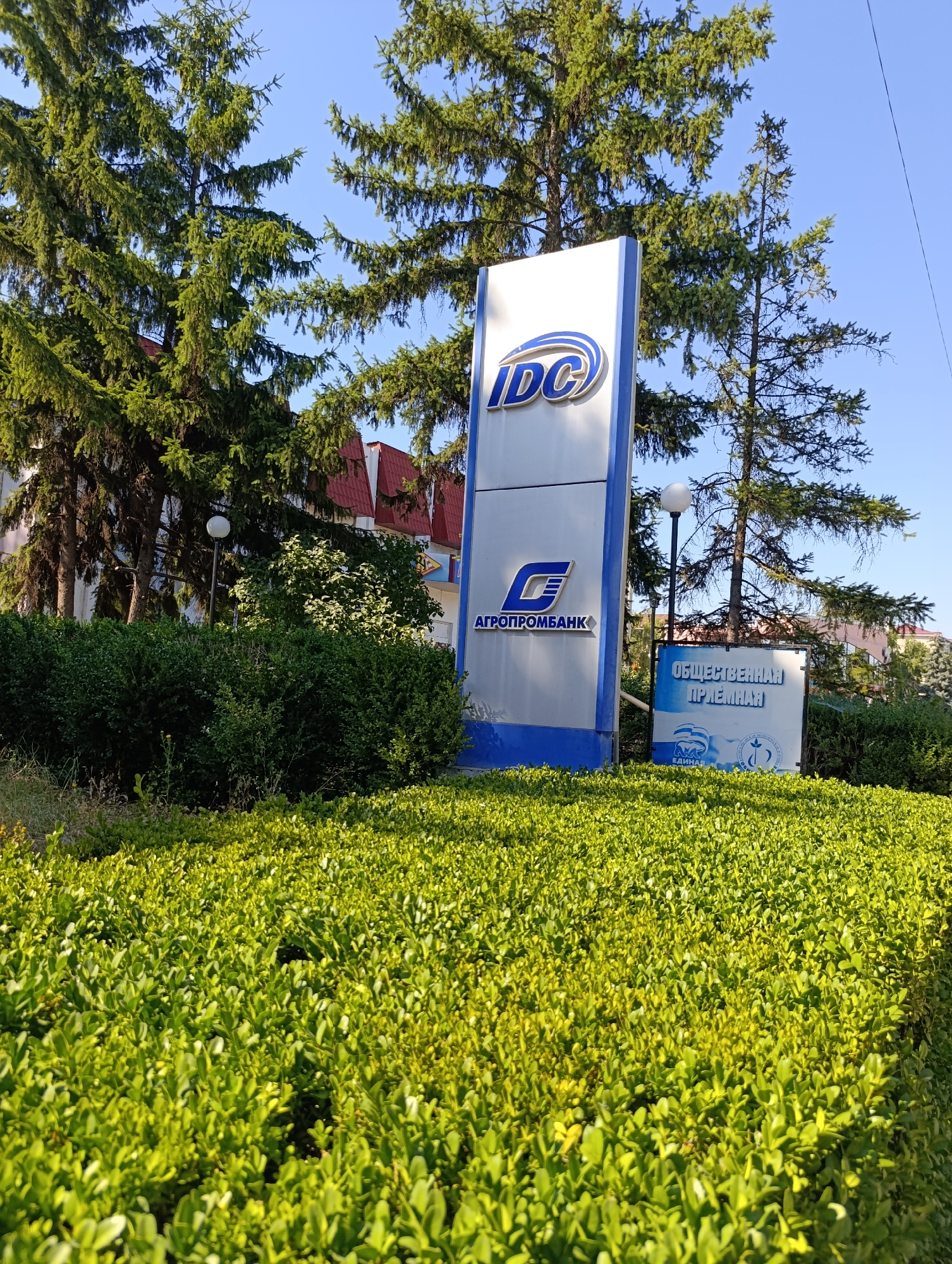 The image shows a signpost outside the town council of Dubăsari, Moldova. The signpost is tall and rectangular, with a blue and white design. The top section features the logo "IDC" in blue lettering within a curved white shape. Below this, the logo for "AGROPROMBANK" is displayed, featuring a stylized blue "E" shape with text underneath. To the right of the main signpost, a smaller, square sign is visible. This sign has a blue and white background with text that appears to say "ОБЩЕСТВЕННАЯ ПРИЕМНАЯ" (Public Reception). Below this text, there are two circular logos, one of which depicts a figure holding a flag, and the other features a stylized globe. The signpost is surrounded by lush green bushes and trees. In the background, buildings with red roofs are visible, along with a clear blue sky. A lamppost with a round white light is also present. The overall scene suggests an exterior view of a public administrative building in a green, well-maintained environment.