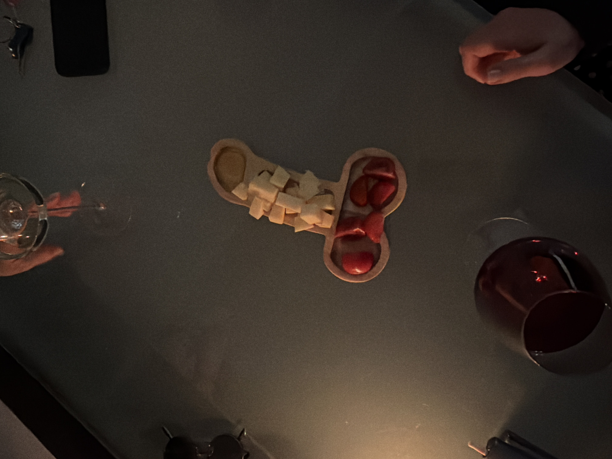 A dimly lit, overhead shot depicts a gathering on a gray surface. At the table's center is a wooden serving dish, its shape suggestive, holding cubes of cheese, strawberries, and a dipping sauce. A half-empty glass of red wine and a nearly empty wine glass are on the right and left respectively. The photo also includes a partially visible hand and a mobile phone. No evidence suggests this image was taken in Moscow, Russia.