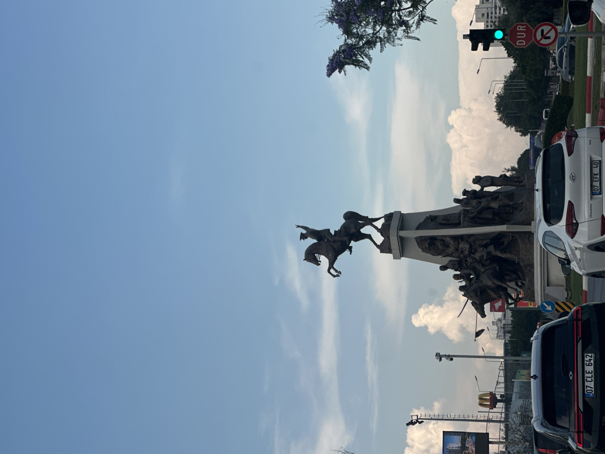 The image depicts a monument with a statue of a man on horseback, along with other figures, in an urban setting under a blue sky with scattered clouds. The monument is located in Konyaaltı, Turkey. In the foreground, two cars are visible. The car on the left is black, and the car on the right is white. A traffic light is illuminated green, and a "DUR" (Stop) sign is posted. To the left of the monument, a McDonald's sign is visible. The overall scene suggests a public space with traffic and urban infrastructure.