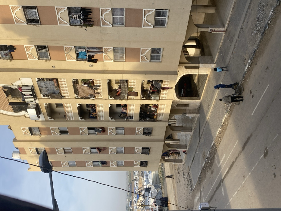 An elevated daytime photograph depicts a multi-story residential building and an adjacent street in Khan Yunis, Palestinian Territory. The prominent structure is a beige-colored apartment building with darker brown vertical accents and white decorative motifs around windows and balconies. It features at least seven visible floors, each with multiple windows and balconies. Many balconies are cluttered with drying laundry, various household items, and improvised tarpaulin or sheet coverings. The ground floor of the building is elevated, supported by a series of arches and columns, creating a shaded underpass or parking area. A water tank is visible on the roof.
In the foreground, an asphalt road with faded white markings runs parallel to the building. A dirt and rubble shoulder separates the road from a paved sidewalk. Three individuals are visible on the sidewalk/street: one adult stands beside a bicycle, another adult in a dark hooded garment is nearby, and a child walks to the right. Several other individuals, including children, are present beneath the elevated ground floor of the building.
To the left of the building and beyond the dirt area, a vast informal settlement composed of numerous makeshift shelters and tents, constructed from various materials, extends into the background. A lone figure walks on a dirt path in front of this camp, and a small group of people are gathered closer to the building. The sky is clear, and the scene is illuminated by natural light, casting shadows from the building and individuals, suggesting morning or late afternoon. A street light fixture and overhead wires are partially visible in the upper left foreground.