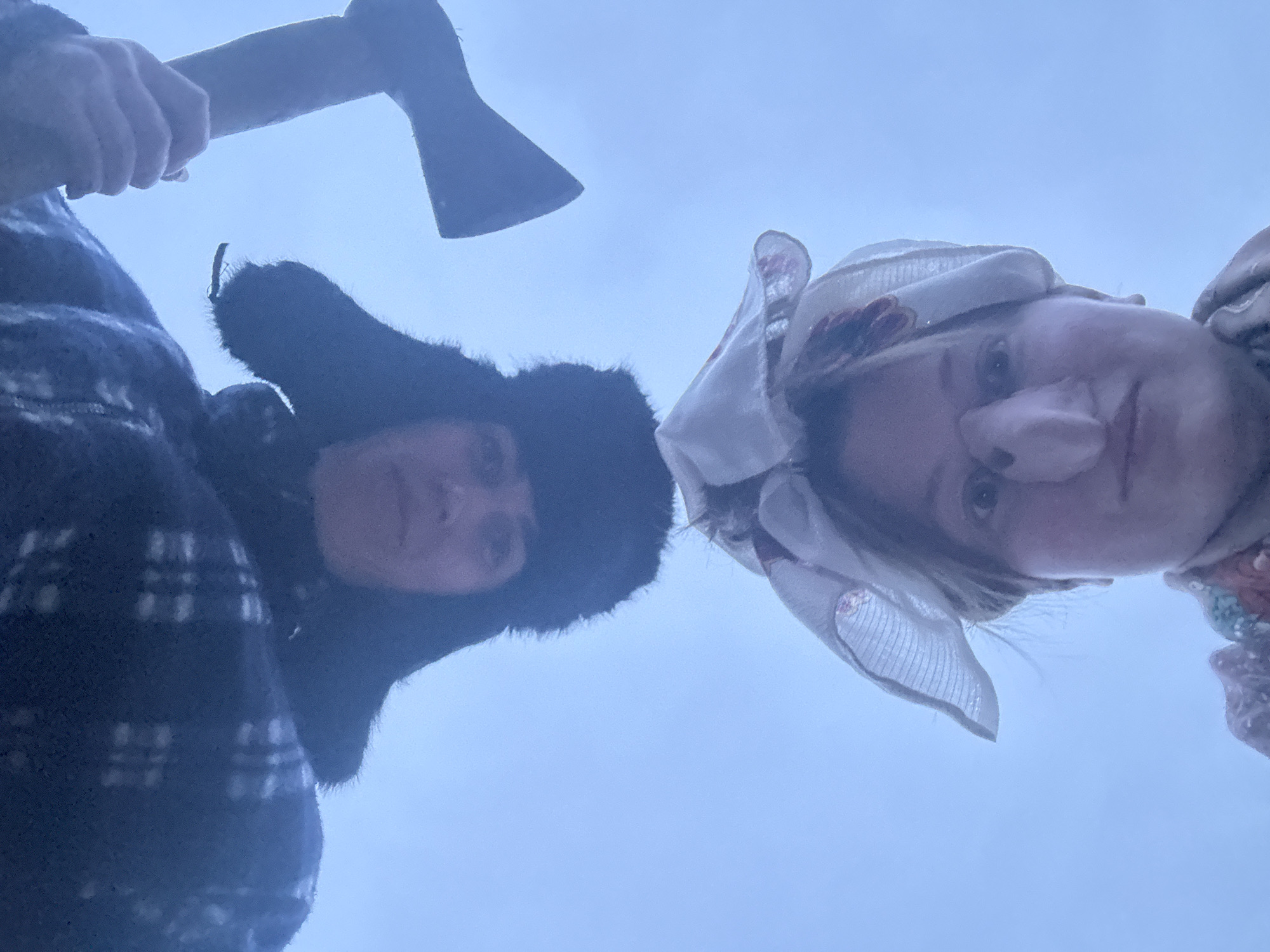 This image, captured in Вассята, Russia, features two individuals looking down at the camera from a low-angle perspective. The background is a uniform light blue, suggesting an overcast sky or a clear, bright sky without discernible features.
On the left, one person is dressed in heavy winter attire, including a dark, possibly plaid or patterned, thick jacket and a dark fur-lined hat, resembling an ushanka. This individual holds an axe with a wooden handle and metallic blade in their right hand, positioned prominently in the upper left of the frame.
On the right, the second person wears a light-colored head covering, appearing to be a patterned headscarf or bonnet, possibly with floral or decorative elements. Their face is partially visible as they look directly down, displaying a neutral to slightly smirking expression.
The heavy clothing worn by both subjects indicates cold weather conditions, consistent with the given location context of Russia. The presence of the axe, combined with the direct gazes, gives the image a dramatic or theatrical quality, suggesting a staged photograph. There is no visible text in the media file.