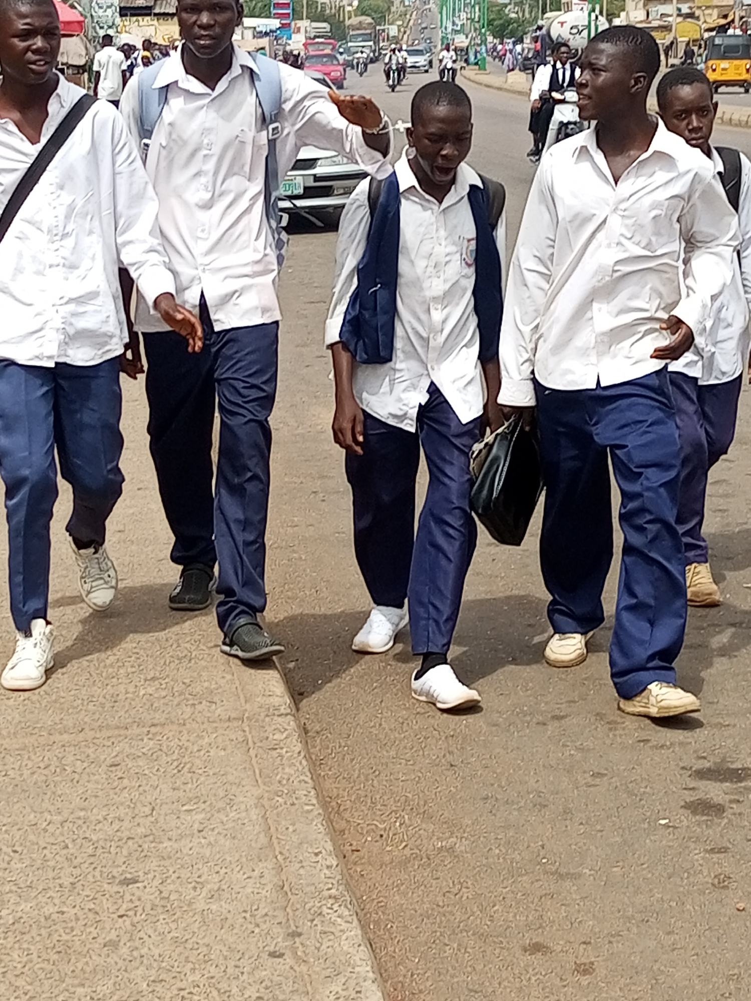In Gombe, Nigeria, five young men in white shirts and blue trousers walk along a paved road. The young men are wearing backpacks, and some are carrying bags. The men are walking in a group, and appear to be students. The road is lined with buildings and vehicles, and there is a lot of activity in the background. The weather appears to be sunny and warm.