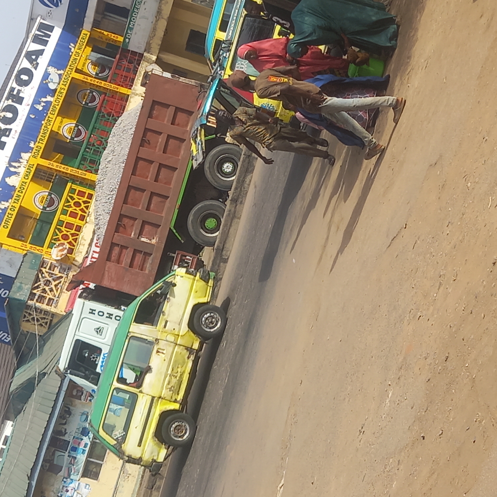 The image captures a street scene in Dadi Riba, Nigeria, featuring a yellow and green minibus parked along the roadside. A brown dump truck loaded with gravel is positioned next to the minibus. Several people are standing near the vehicles. In the background, signs for "ROFOAM" and "OFFICE OF YAN DOYA CHAPEL ROAD TRANSPORT EMPLOYER'S ASSOCIATION OF NIGERIA" are visible. The street surface is dusty.