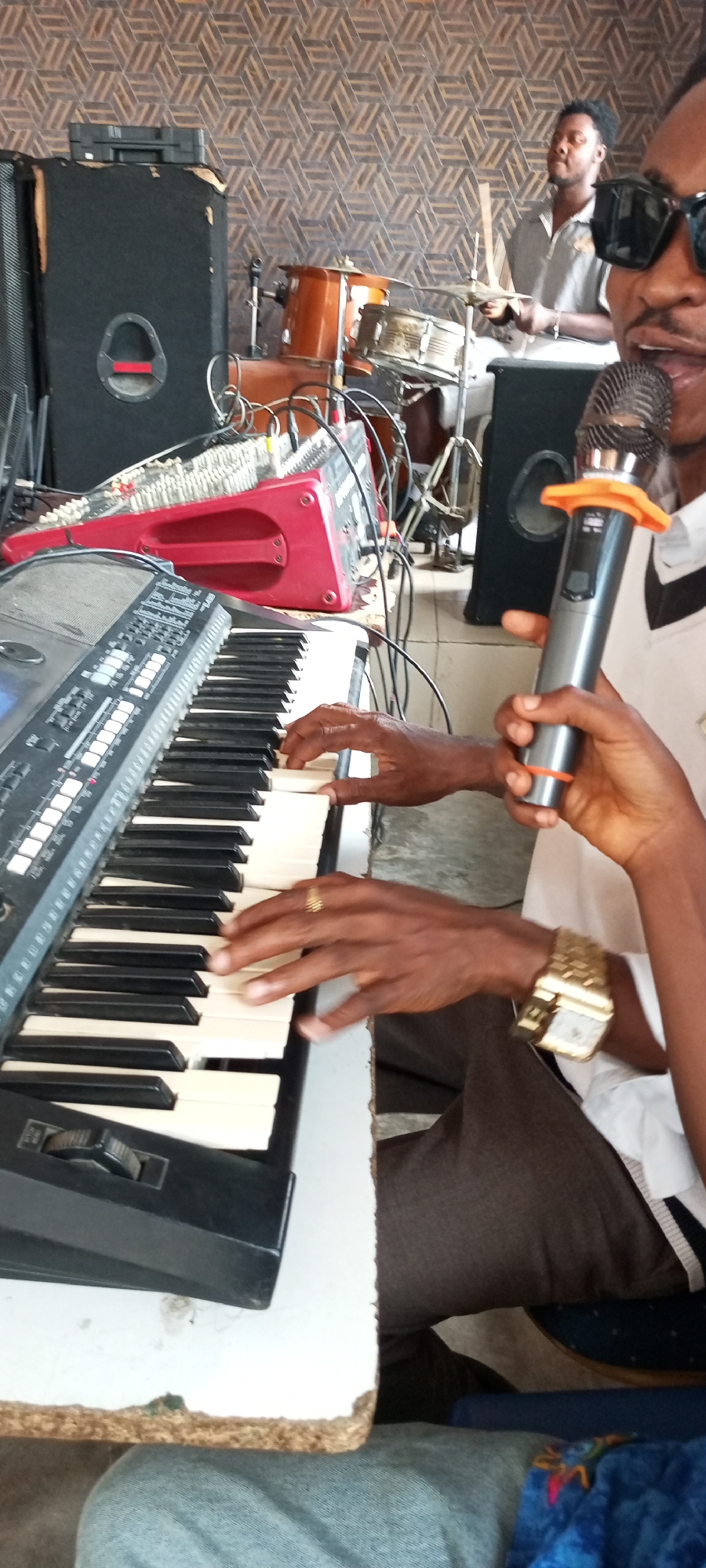 The image shows a musical performance taking place indoors, possibly in a church or a recording studio, in Lokoja, Nigeria. In the foreground, a man is playing a keyboard and holding a microphone. He wears a gold watch and a gold ring. Behind him is a red mixing console and a large black speaker. Further in the background, another man plays drums. The room has a patterned wall, resembling either a wallpaper or a decorated panel. The scene seems to depict a live music session.
