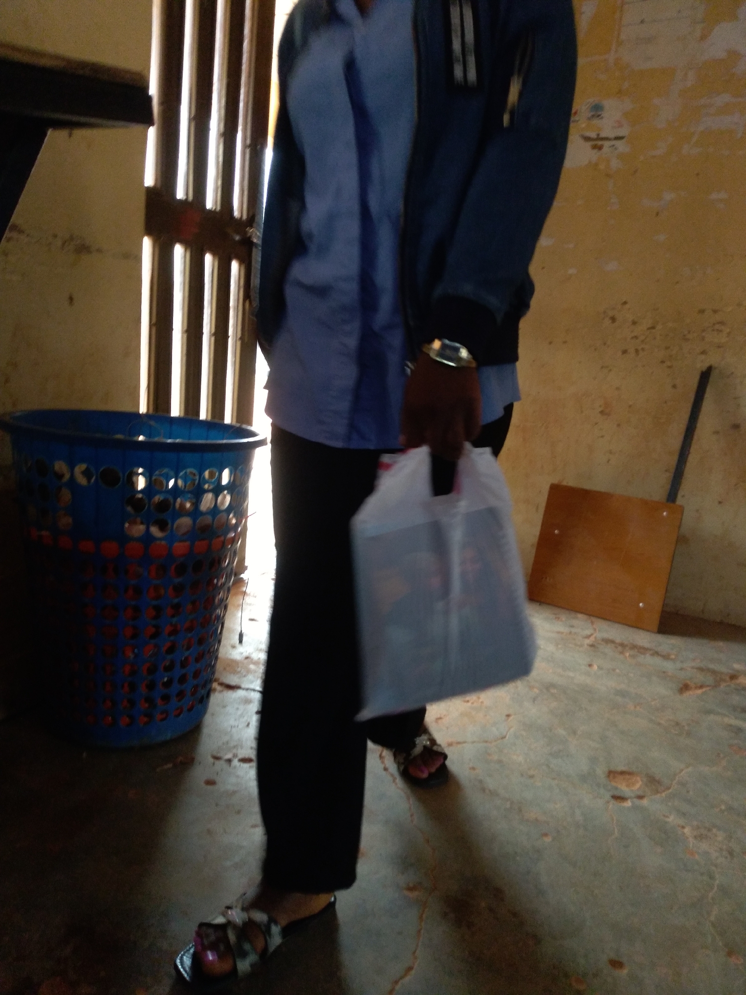 A medium shot captures a person standing in what appears to be a dimly lit indoor space with a dirt floor. The person is wearing dark pants, a blue collared shirt, and a dark jacket. They are holding a clear plastic bag filled with items in their right hand. A large blue plastic basket with a perforated design is visible to the left of the person, containing red objects. The background features a wall with peeling paint and a wooden object leaning against it. The scene suggests a functional, possibly utilitarian, environment. The specific location in Jos, Nigeria is not discernible from the visual information.