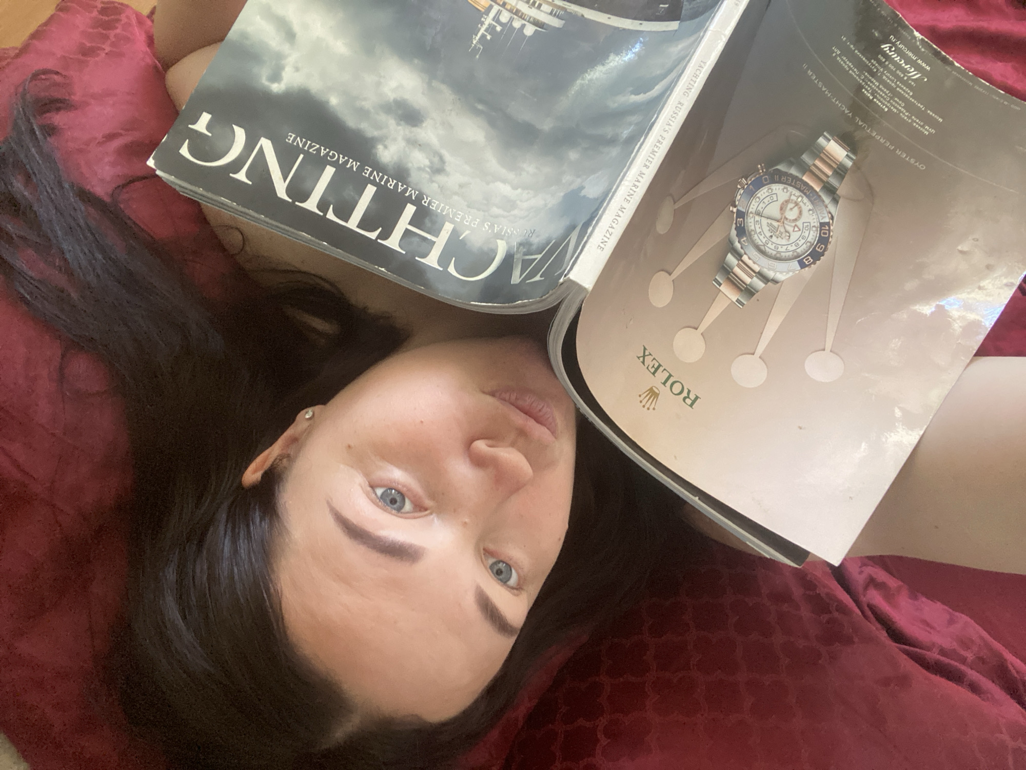 A person is lying on a red patterned fabric, obscured by two magazines. The magazine on the left is titled "YACHTING RUSSIA'S PREMIER MARINE MAGAZINE". The magazine on the right displays a Rolex Oyster Perpetual Yacht-Master II watch. The person's face is visible between the magazines, with dark hair, blue eyes, and a pierced ear. According to text in the magazine, the watch is available in the Russian city of Sochi at the Grand Marina.